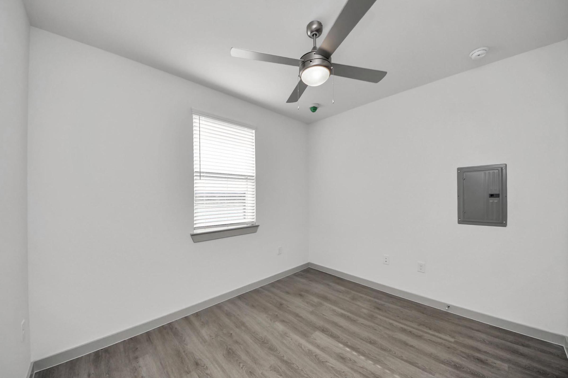 An empty room with light gray walls, a ceiling fan with a light fixture, and a window with white blinds. The floor is a light wood laminate, and there is an electrical panel mounted on one wall. The overall feel is modern and minimalistic, with abundant natural light.