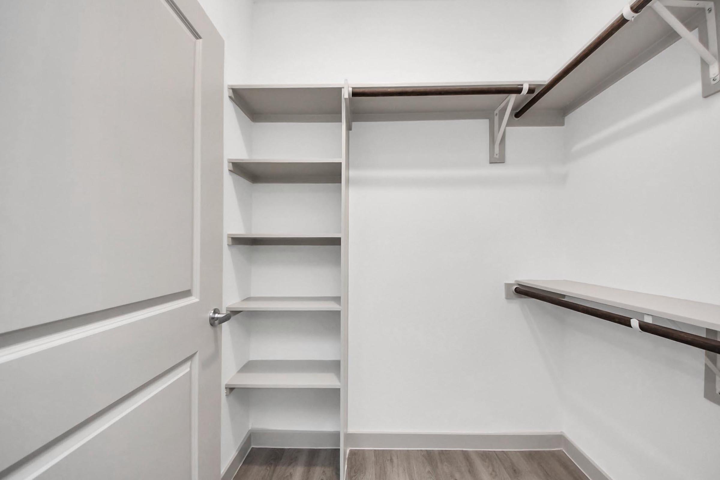 An empty closet interior featuring a light-colored wall with wooden shelves and hanging rods. The floor is a light wood laminate, and a closed door is visible on the left side. The space is well-lit and organized, with a minimalistic design.