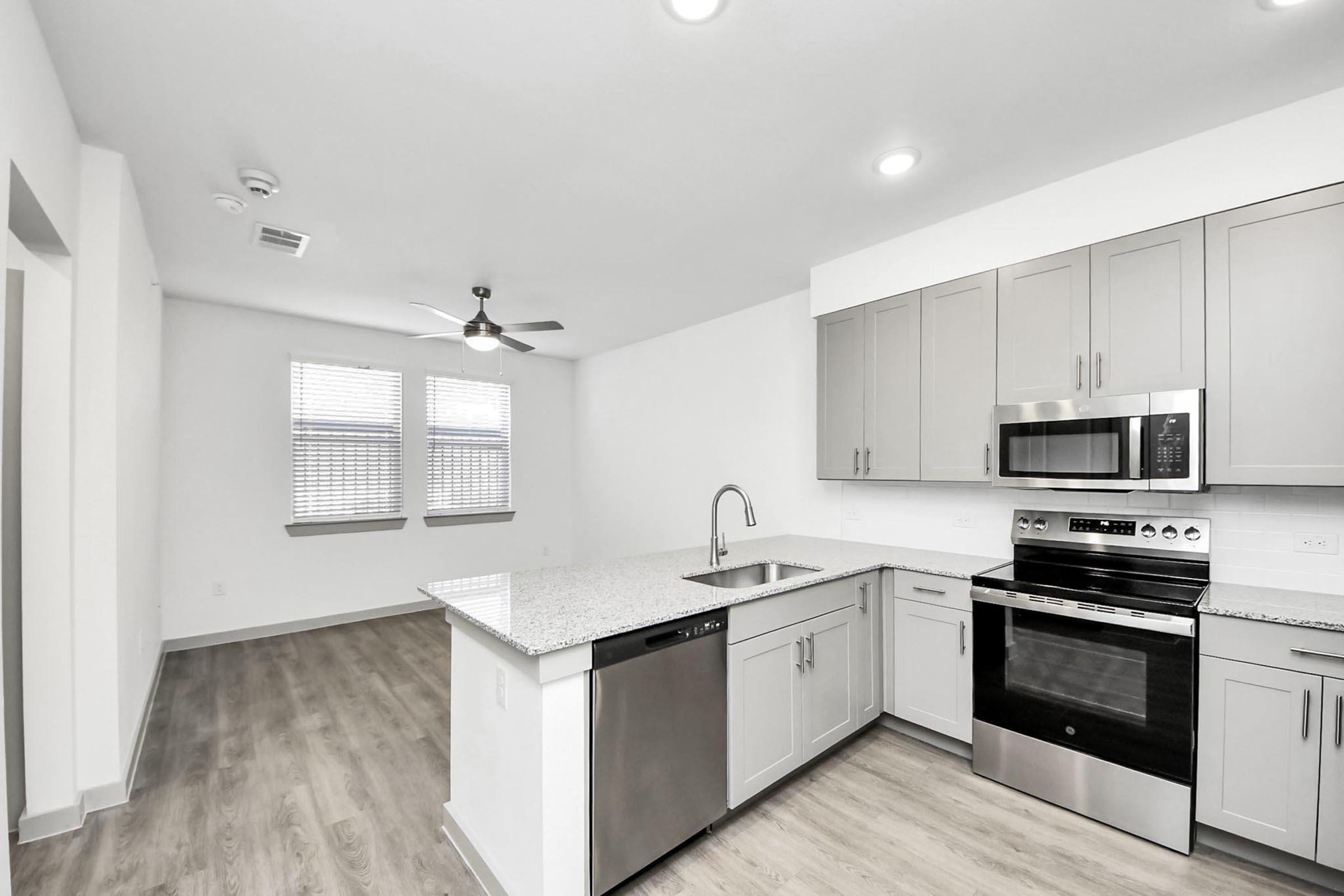 Modern kitchen featuring granite countertops, stainless steel appliances including an oven and microwave, a dishwasher, and a sleek sink. The space has light-colored cabinets and a ceiling fan, with large windows allowing natural light into the adjacent living area.
