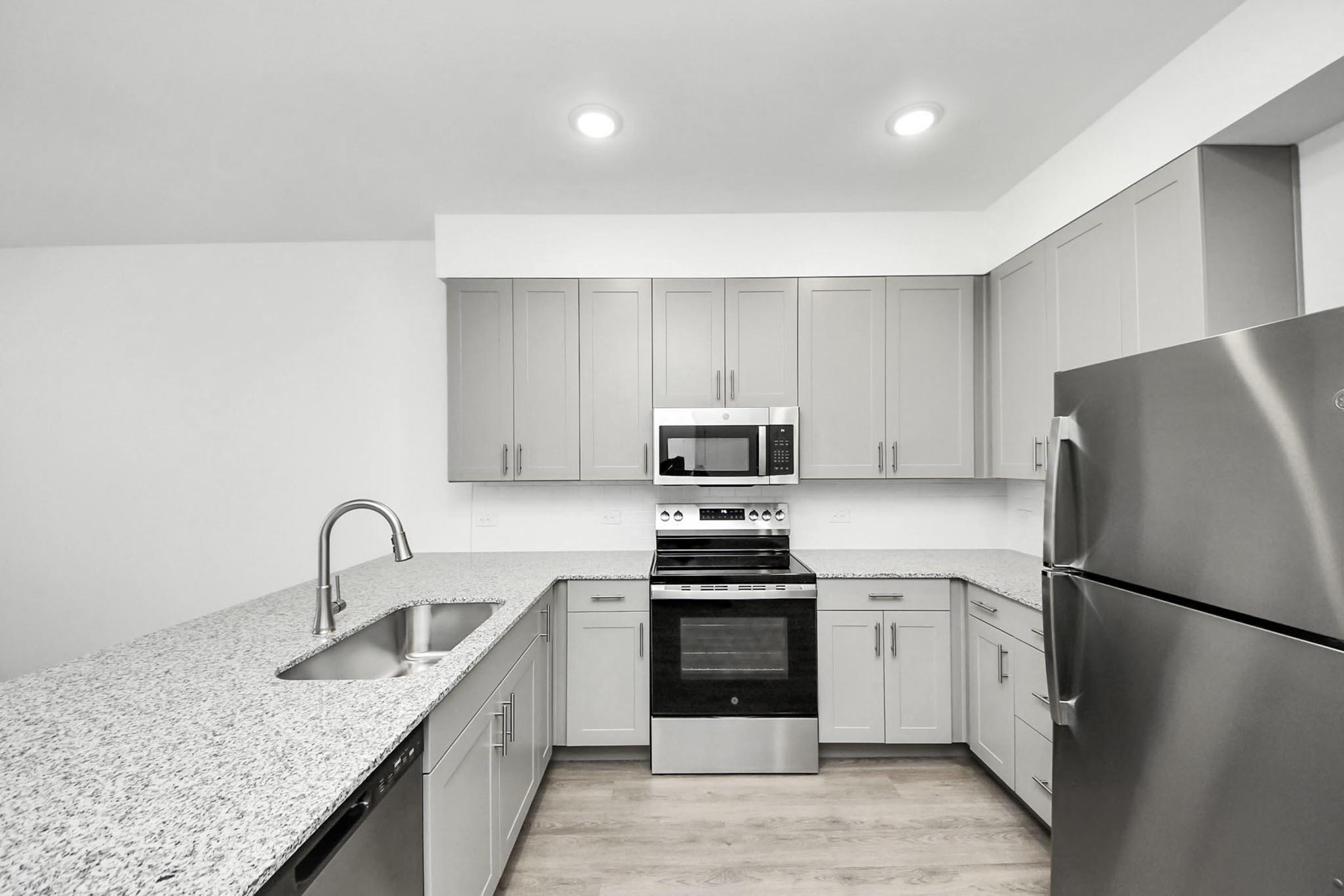 A modern kitchen featuring light gray cabinets, a stainless steel refrigerator, a microwave above the oven, and a granite countertop with a double sink. The space is well-lit with recessed lighting and has a minimalist design, emphasizing clean lines and a contemporary aesthetic.