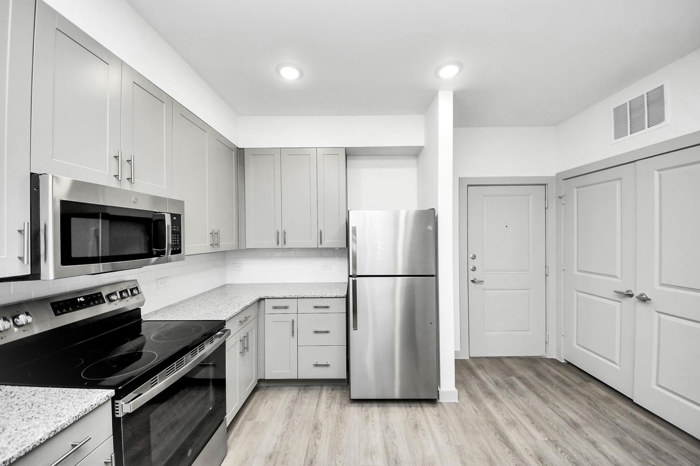 Modern kitchen featuring light gray cabinetry, a stainless steel refrigerator, and a microwave above the stove. Countertops are made of a light-colored granite, and the space has warm wood-like flooring. The room also includes a view of a doorway and an adjacent wall with a door.