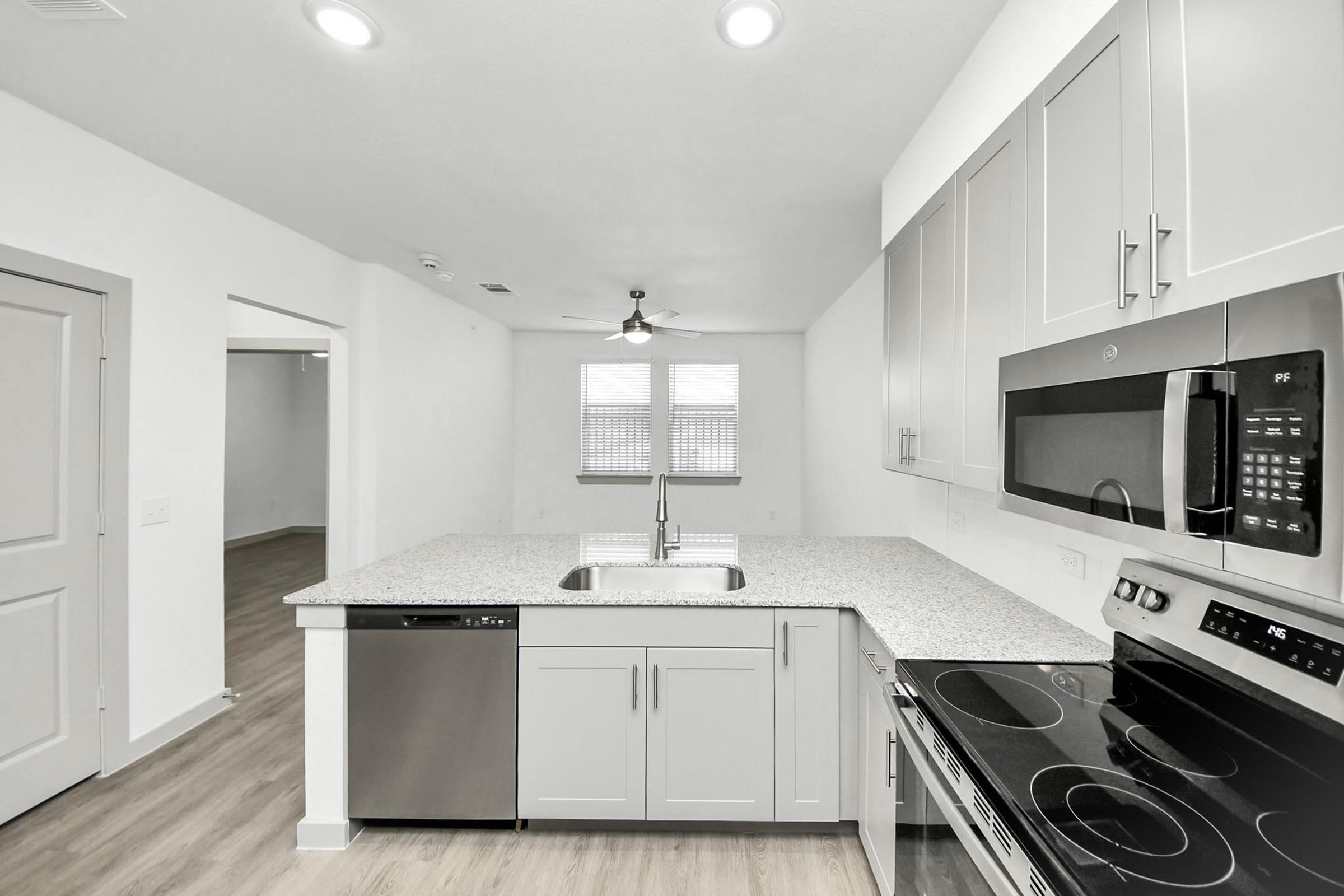 Modern kitchen with light gray cabinetry, a large granite countertop, stainless steel appliances including a microwave, oven, and dishwasher. There’s a sink in the center of the island, and natural light streams through windows. The adjacent area leads to a spacious living room.