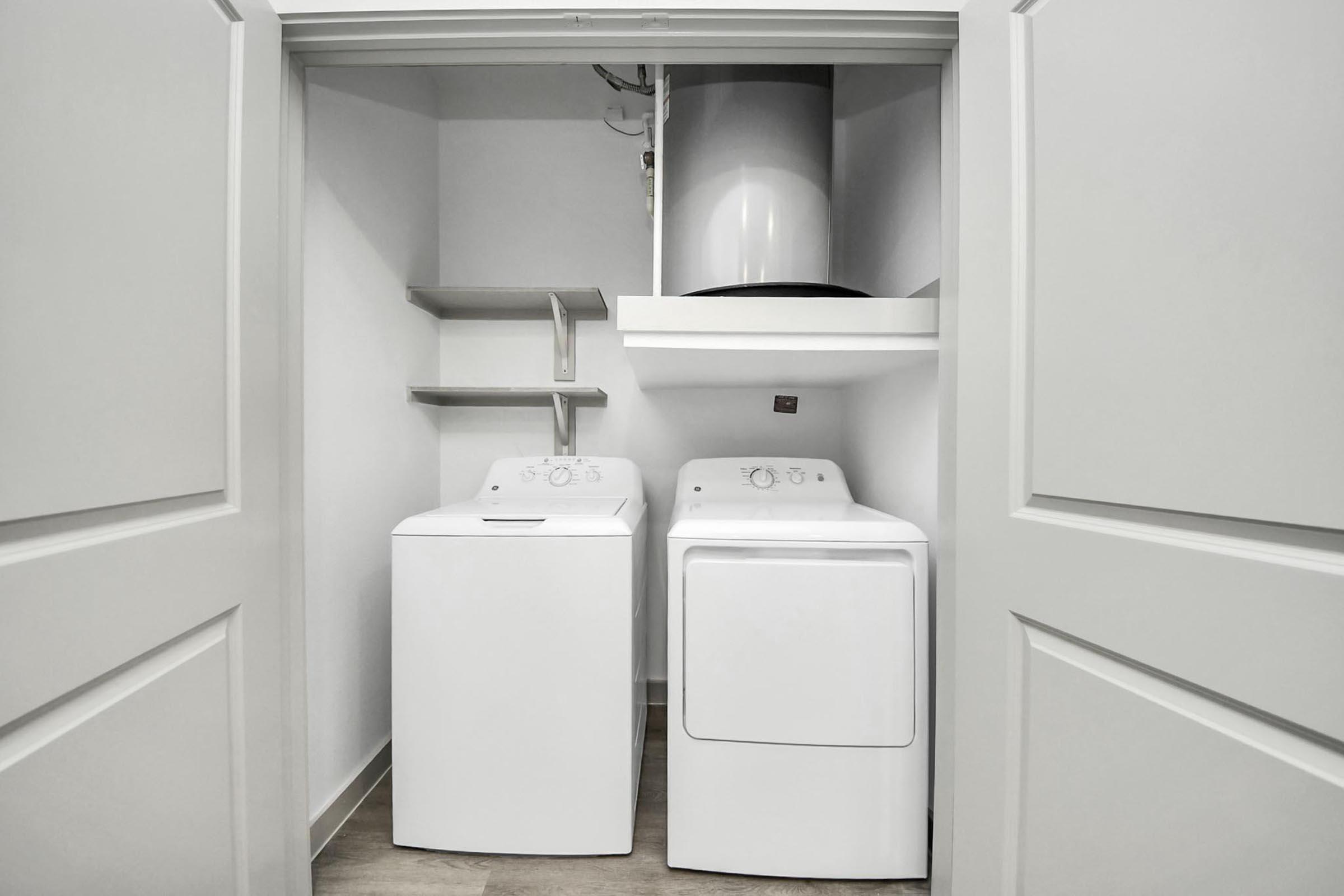 A small laundry room featuring a washing machine and a dryer side by side. The laundry appliances are white, and there is a shelf above the dryer. The doors to the laundry area are open, revealing a clean and organized space with light-colored walls and a light floor.