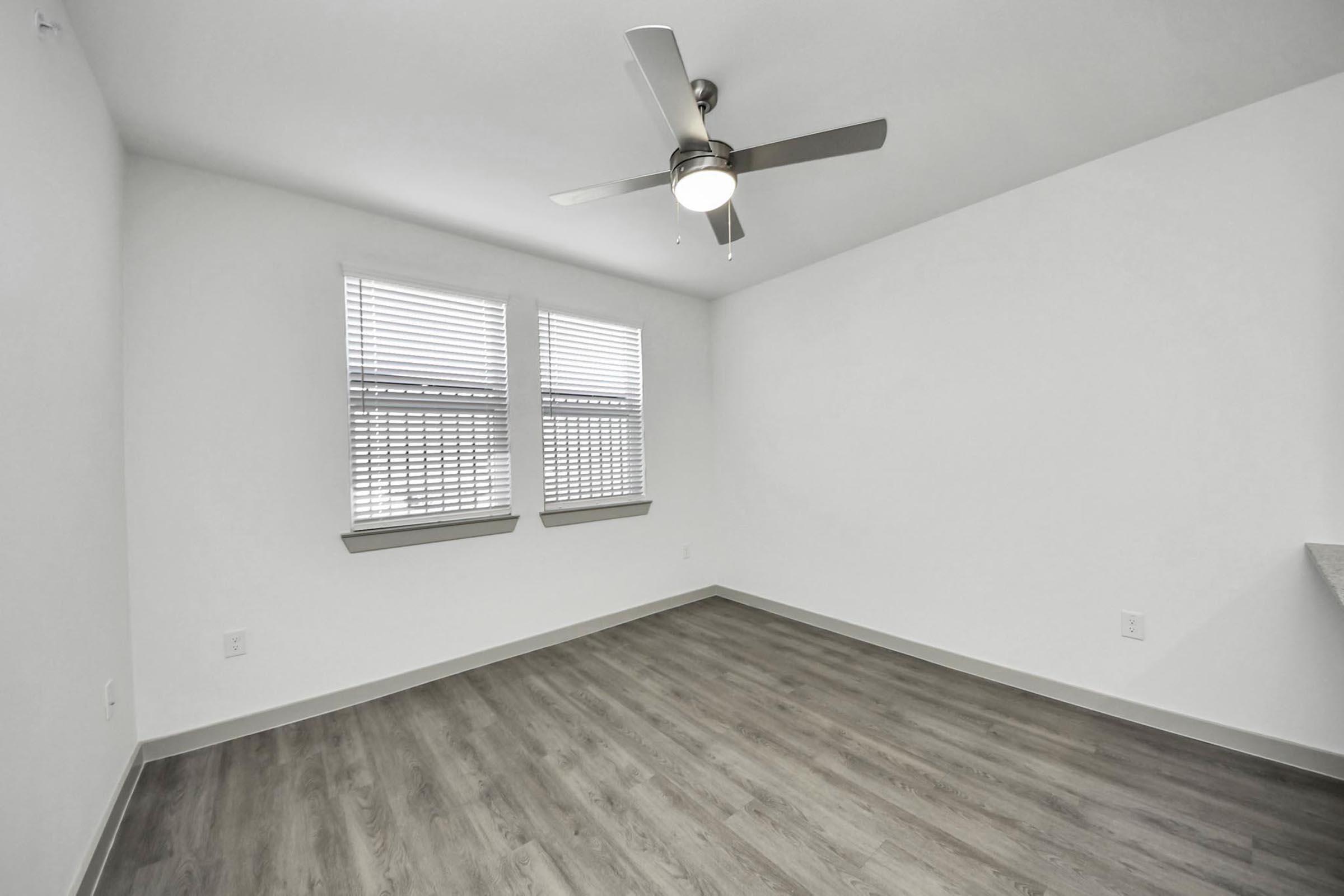 Bright and empty room with light-colored walls, two windows with blinds, a ceiling fan, and wood-like flooring. The space is minimalistic and modern, featuring a clean design without furnishings.