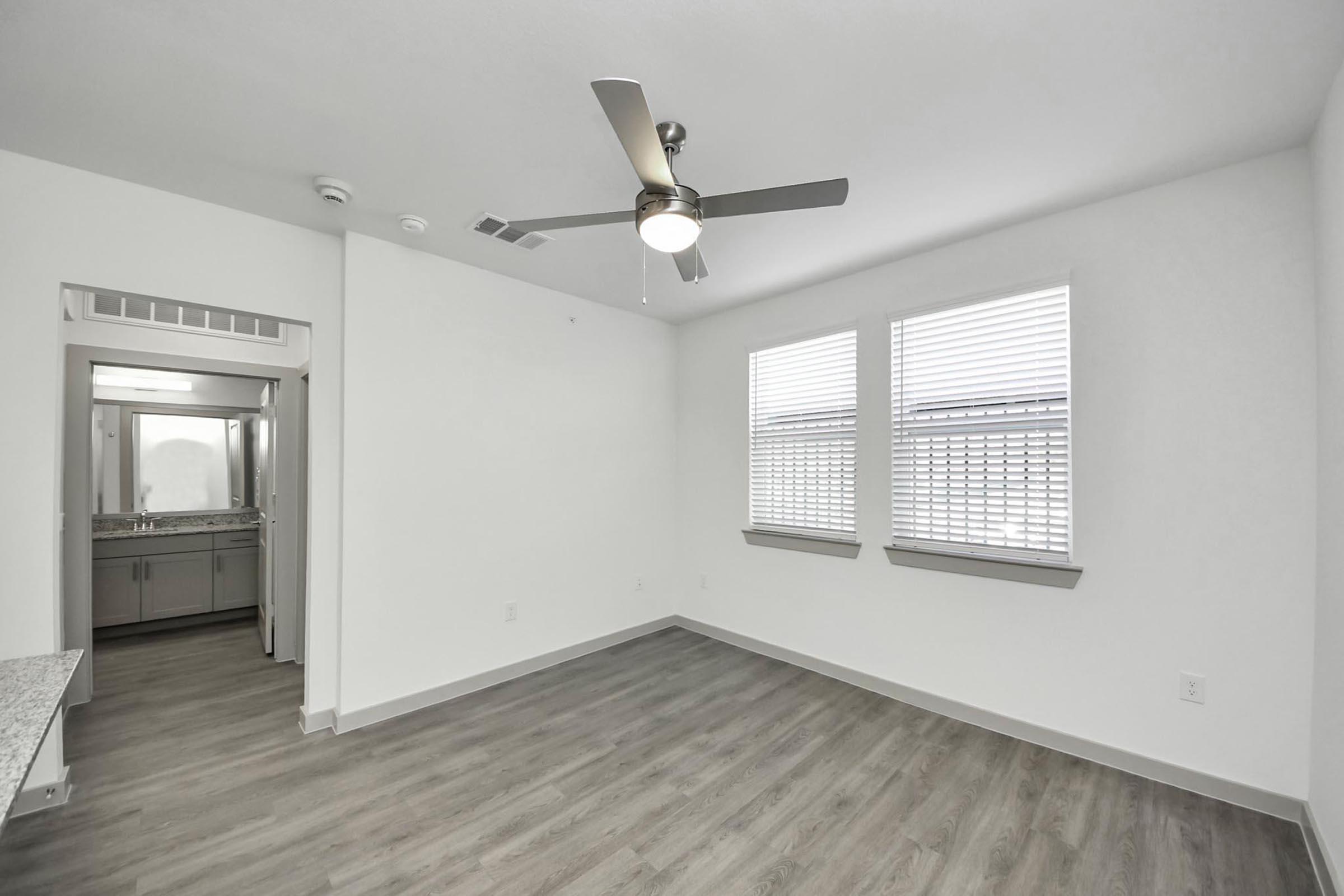 Spacious interior of a modern room featuring large windows with blinds, a ceiling fan, and light-colored walls. A light-colored wooden floor completes the contemporary look. A glimpse of a bathroom is visible through a doorway, showcasing a clean and organized design.