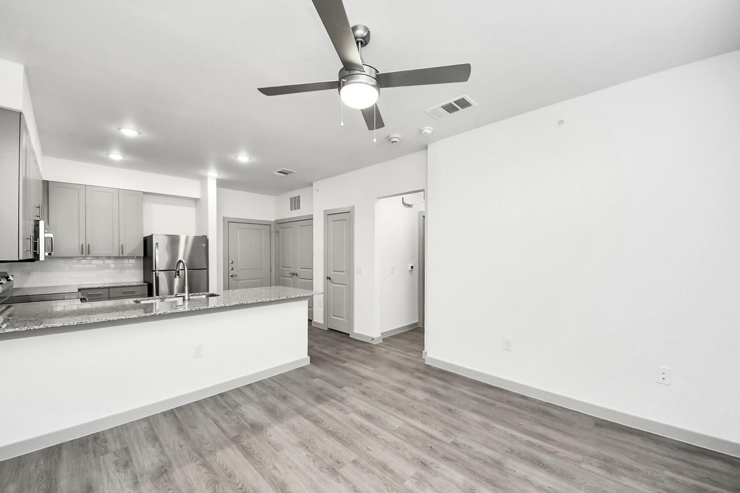 A modern, open-concept apartment interior featuring a kitchen with stainless steel appliances, a large island, and a living area with laminate flooring. There is a ceiling fan, light-colored walls, and a doorway leading to another room. The space is well-lit and minimalistic in style.