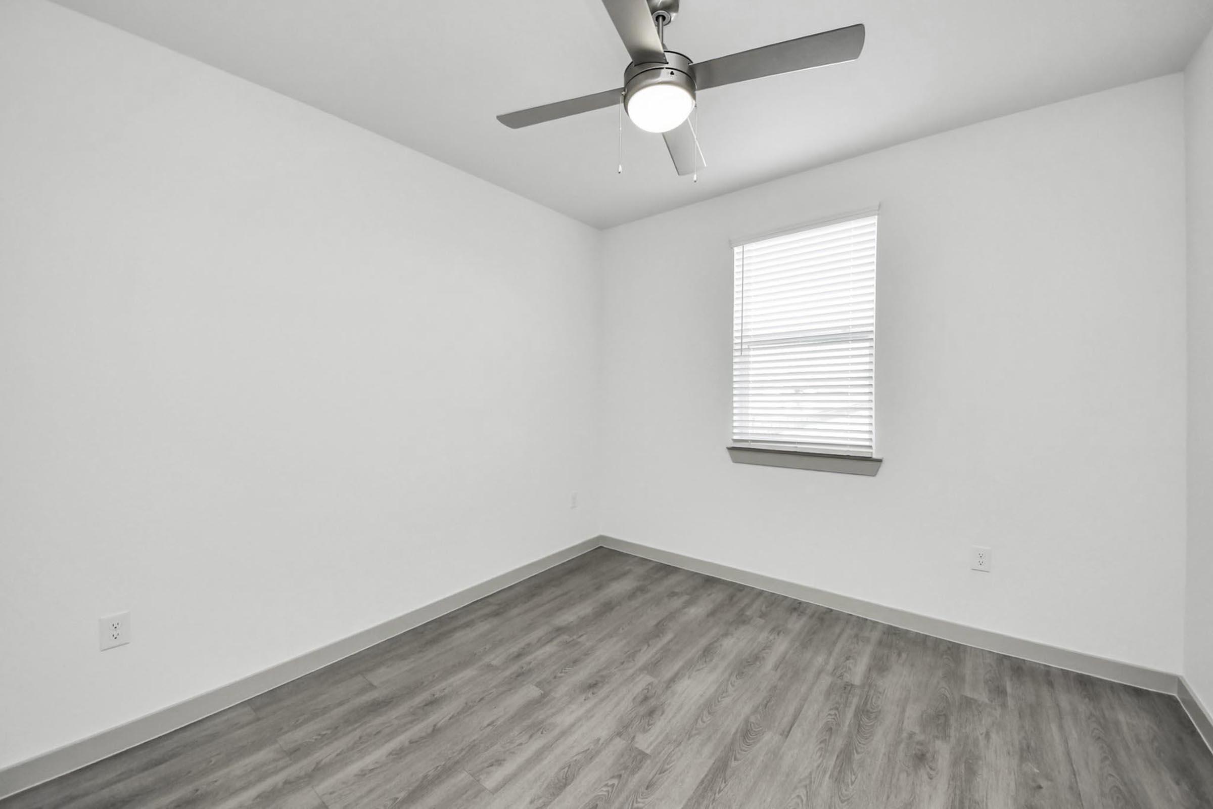 Empty room with light gray laminate flooring, a ceiling fan, and a window covered with white blinds. The walls are painted white, and there's a small ledge below the window. At the corner, the room features a neutral color scheme, giving it a bright and airy atmosphere.