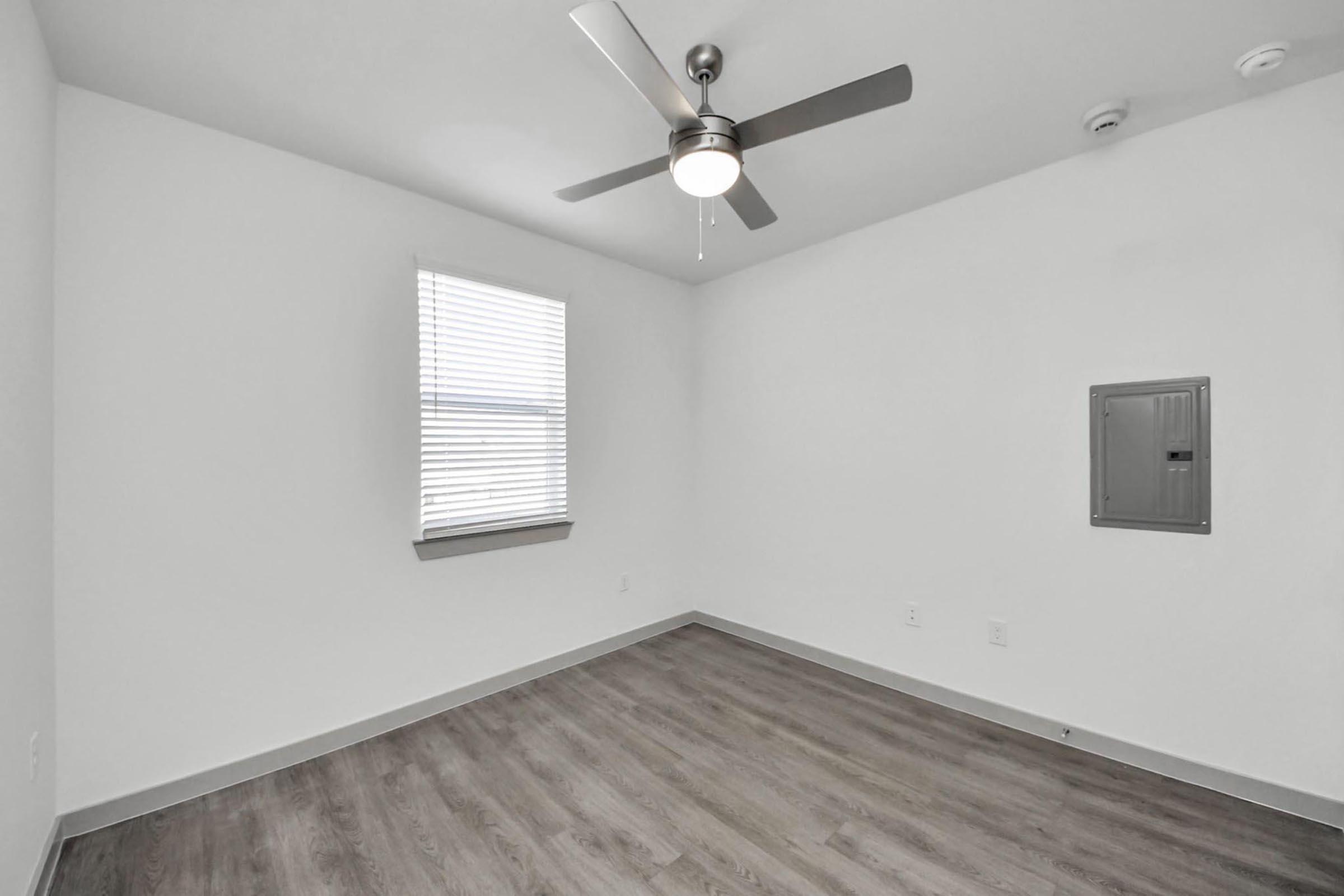 A minimalist interior of a bright room featuring a light-colored wall, a window with blinds, and a ceiling fan. The floor has light wooden planks, and there is a small electrical panel on one wall. The room is empty, creating a spacious feel.