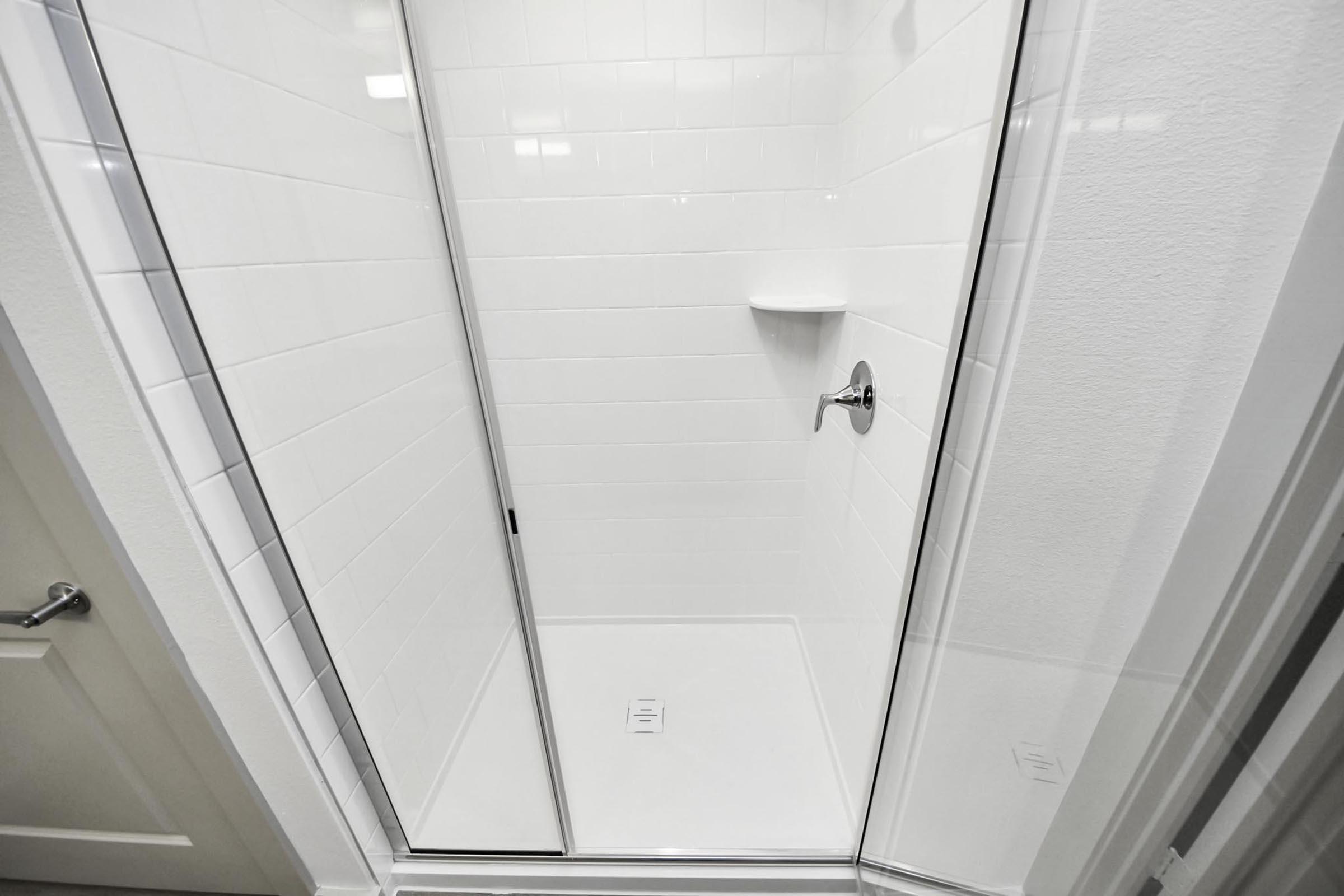 A clean, modern shower stall featuring clear glass doors and white tiled walls. The shower has a simple design with a built-in shelf, a wall-mounted faucet, and a drain on the floor. The overall appearance is bright and minimalistic, ideal for a contemporary bathroom setting.