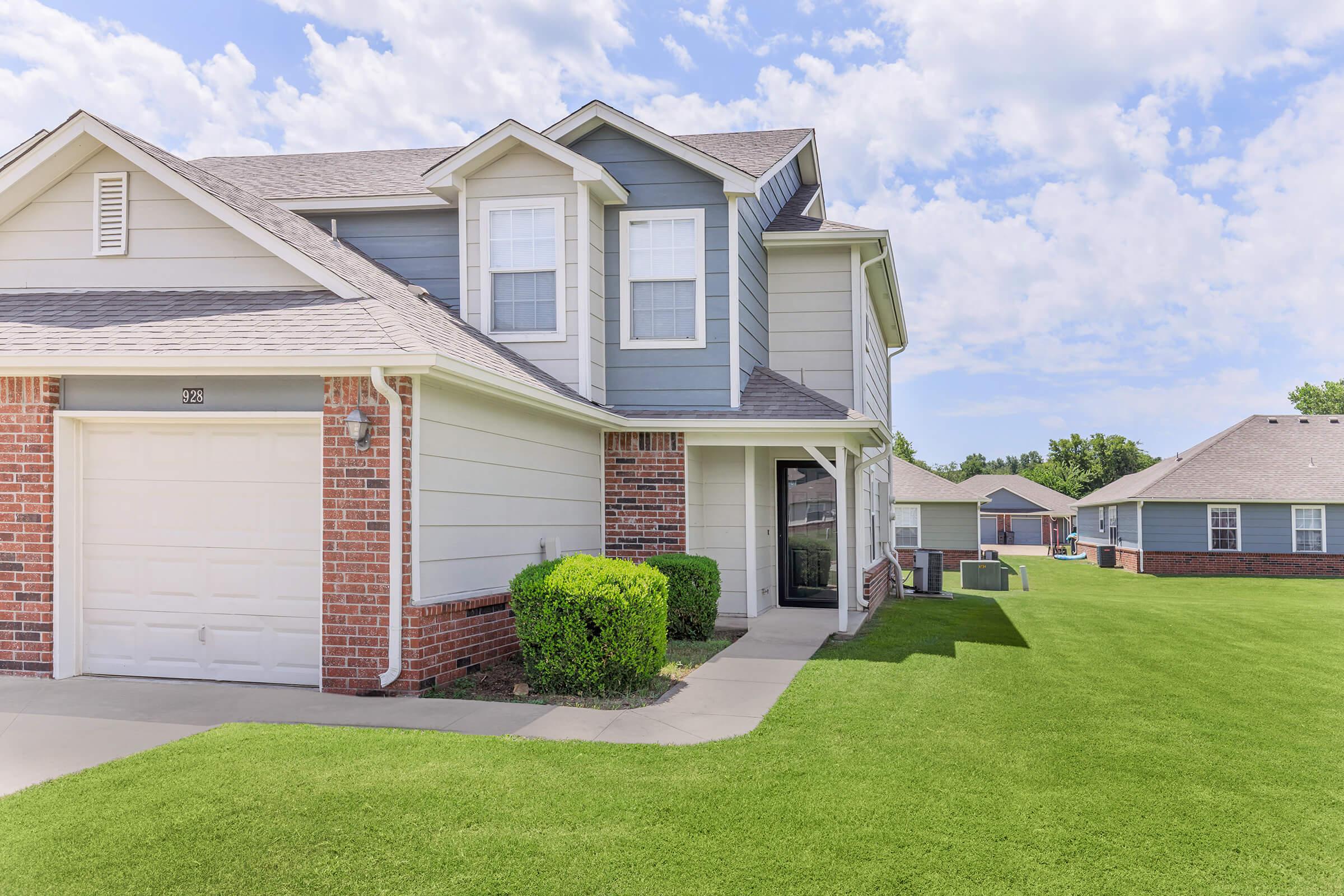 A two-story house with a light gray exterior and brick accents, featuring a garage and an entrance. The well-maintained lawn is neatly trimmed, and the sky is partly cloudy. Other similar houses can be seen in the background, creating a suburban neighborhood atmosphere.