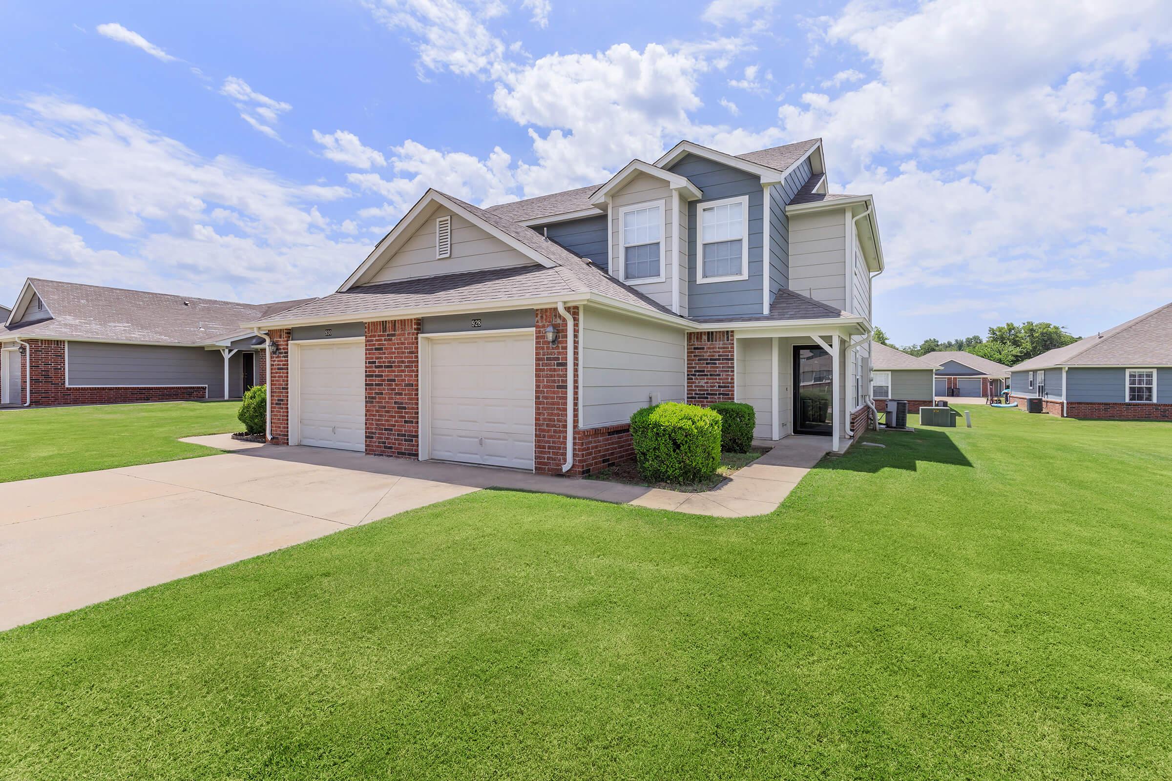 Two-story residential house with a light gray exterior and brick accents, featuring a front porch and two garages. The well-maintained lawn is green and expansive, with a blue sky and scattered clouds in the background.