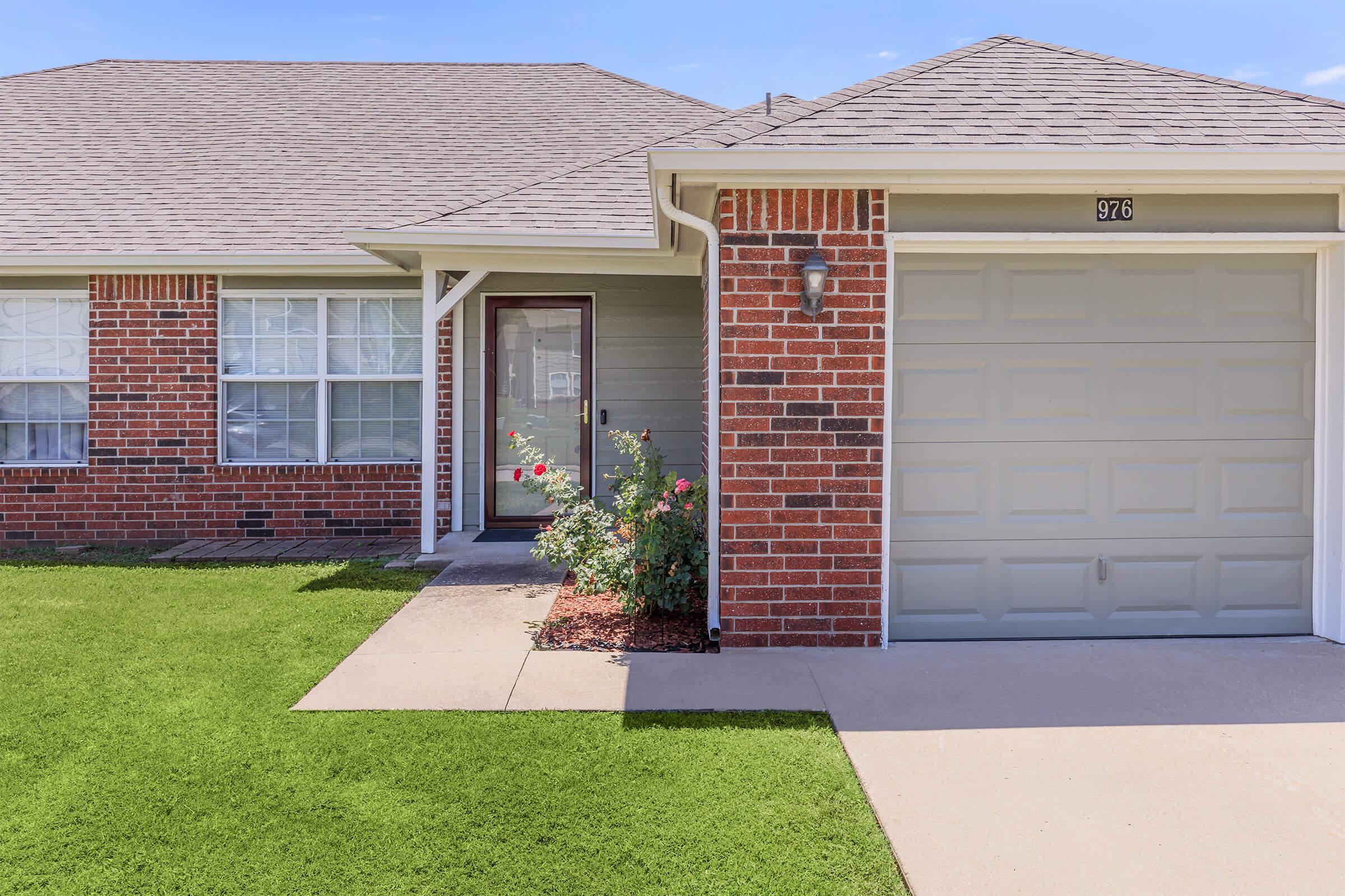 A well-maintained single-story brick house with a light gray garage door. The front yard features neatly trimmed grass and a small garden with blooming rose bushes near the entrance. The house has multiple windows, and the roof is sloped. The overall appearance is inviting and well-kept.