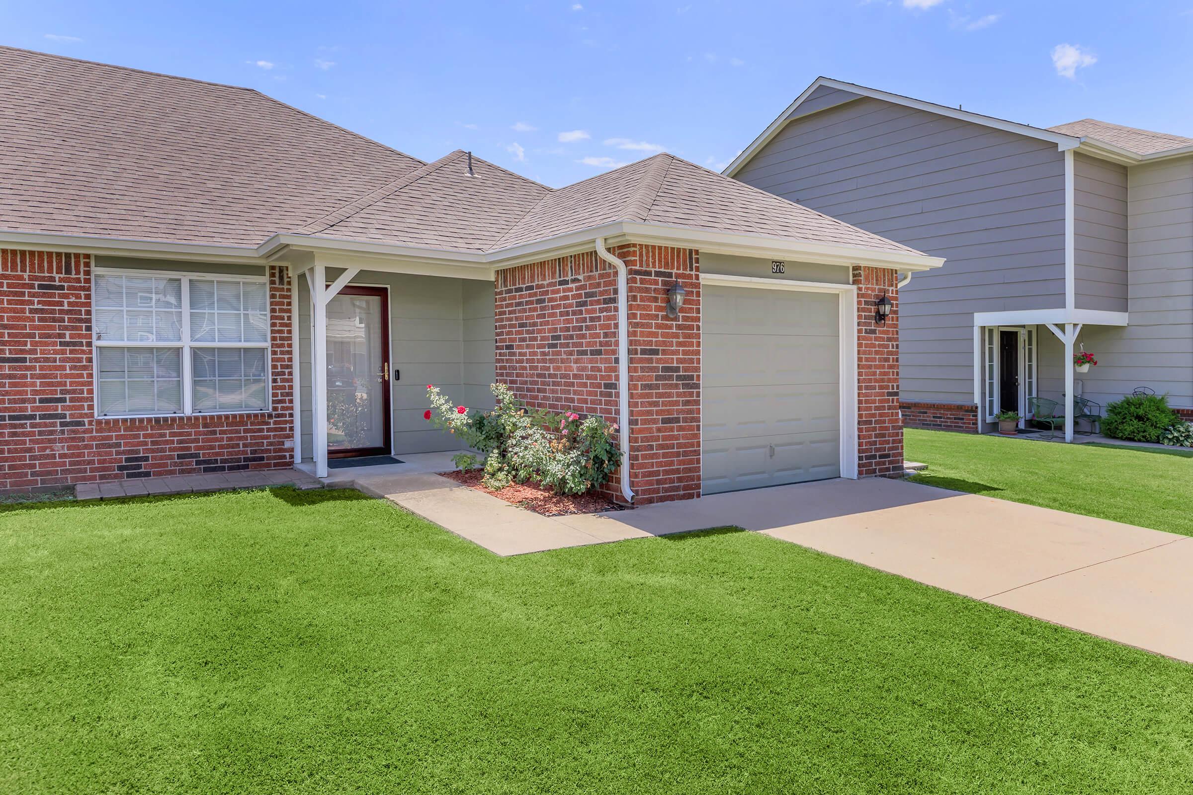 A well-maintained suburban home featuring brick exterior, a covered porch, and a single-car garage. The front yard has neatly trimmed grass and a few flower bushes, surrounded by a clear blue sky. Adjacent homes are visible in the background, showcasing similar architectural styles.