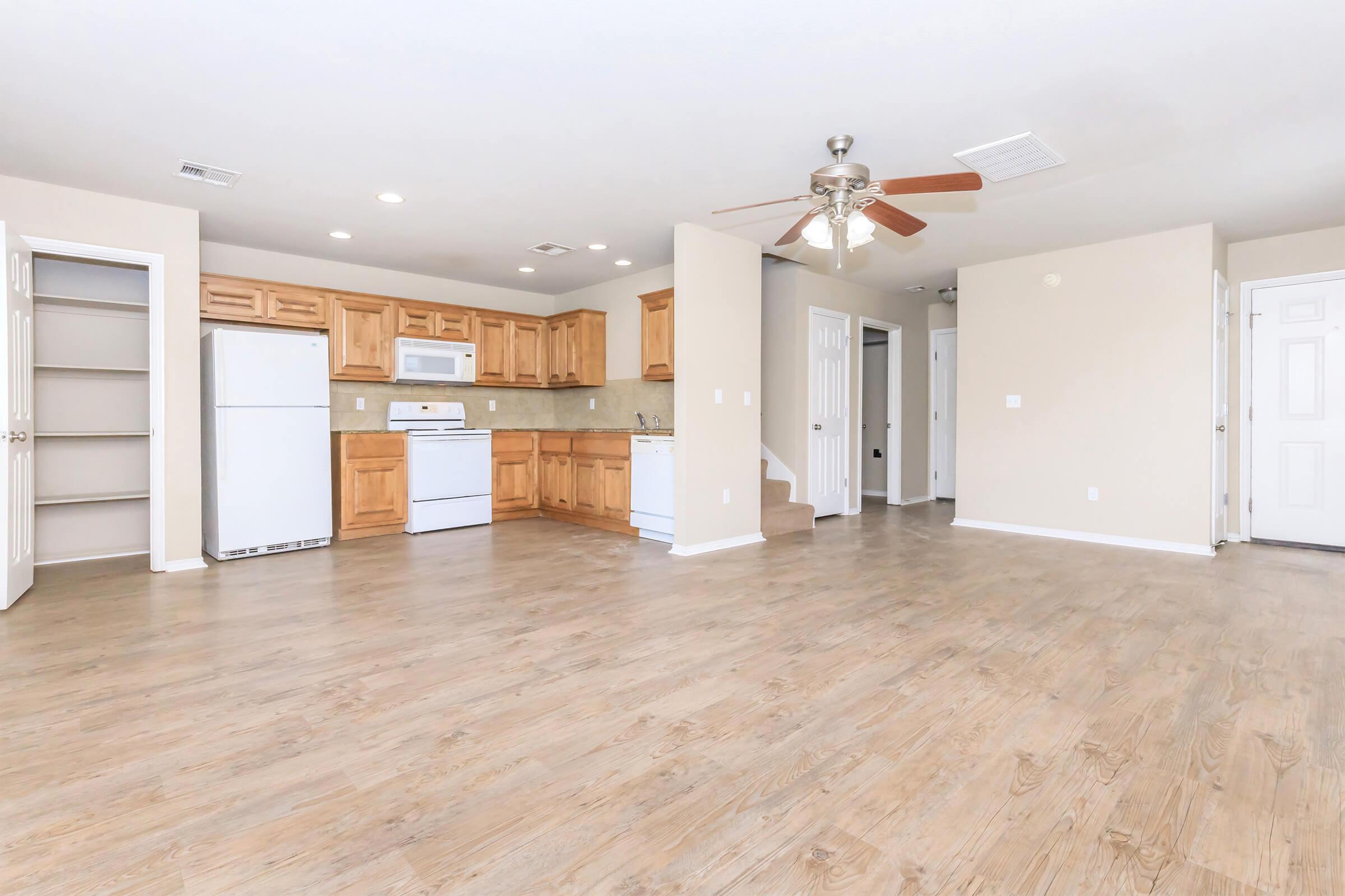 Spacious open-concept living area featuring a modern kitchen with wooden cabinetry, white appliances, and a ceiling fan. Light-colored walls and wood-like flooring create a warm atmosphere. Includes a pantry space and doors leading to other rooms for easy access.