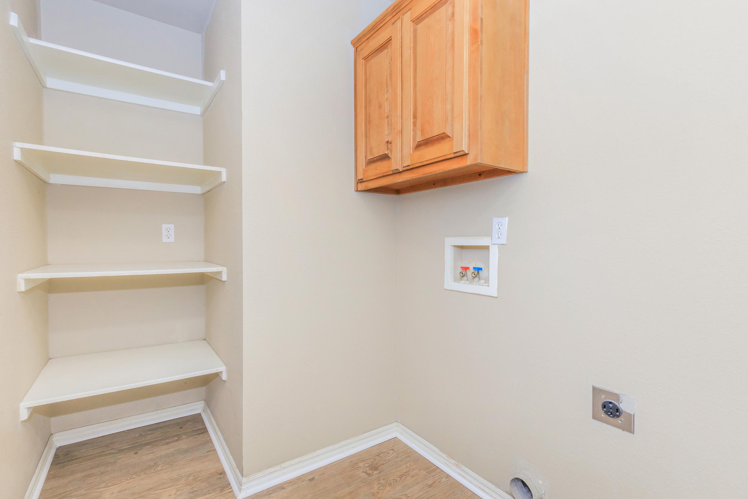 A small, light-colored room featuring a built-in shelf unit on the left side, a wooden cabinet mounted on the wall, and a small wall cutout with two electrical outlets. The floor is wood-like, and the walls are painted a neutral shade. Overall, the space looks tidy and organized.