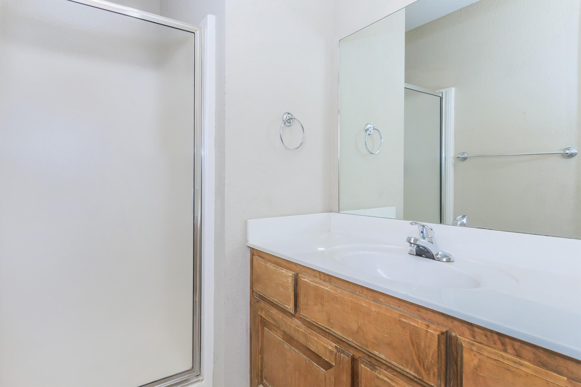 A clean and modern bathroom featuring a white sink with a chrome faucet, a wooden cabinet below, and a large mirror above. On the right, a shower stall with a frosted glass door is visible, and the walls are painted in a light neutral color.