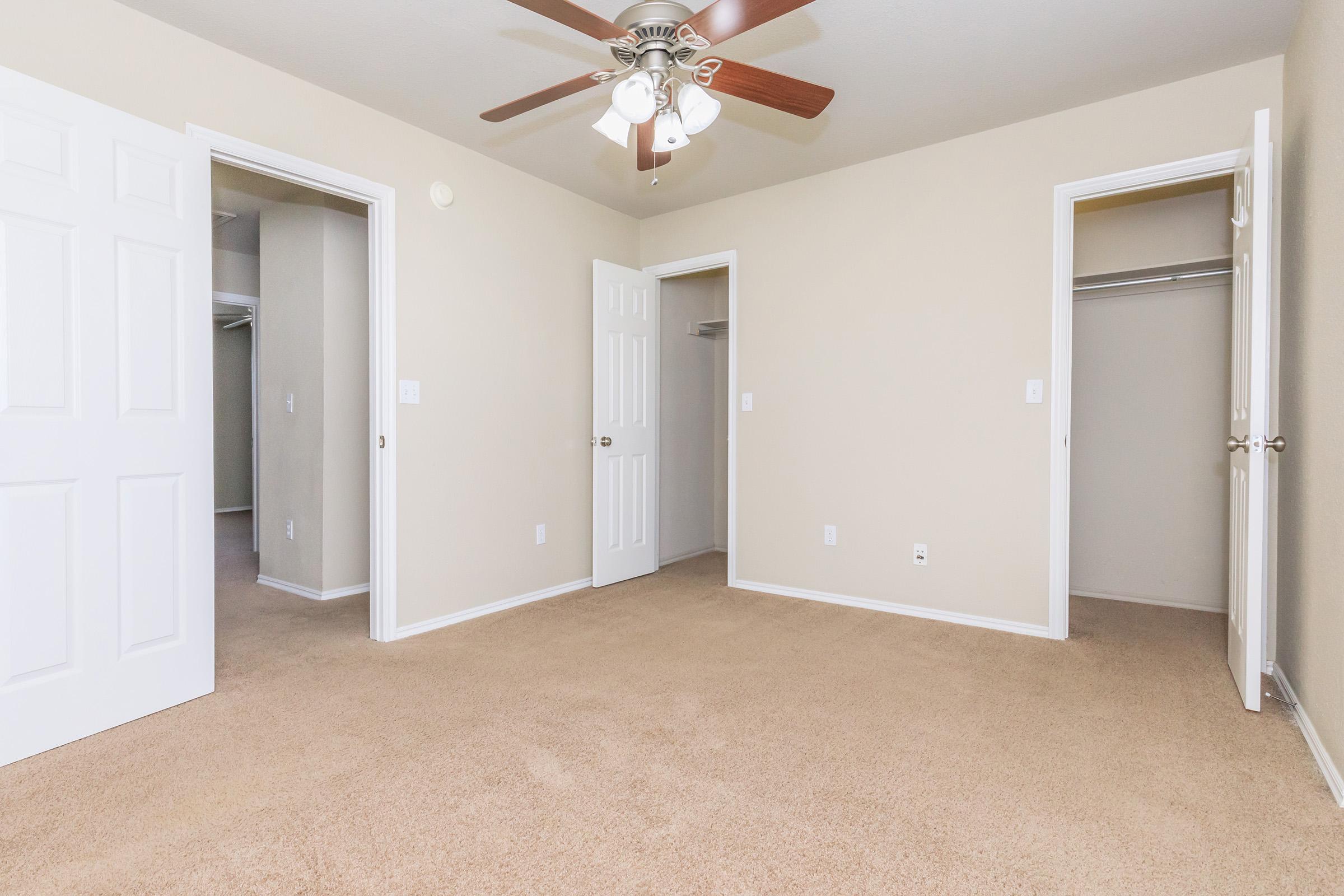 A spacious, neutral-colored bedroom featuring a ceiling fan, light fixtures, and two open doors leading to closets. The carpeted floor adds warmth, and the walls are painted in a light beige color, creating a bright and inviting atmosphere.