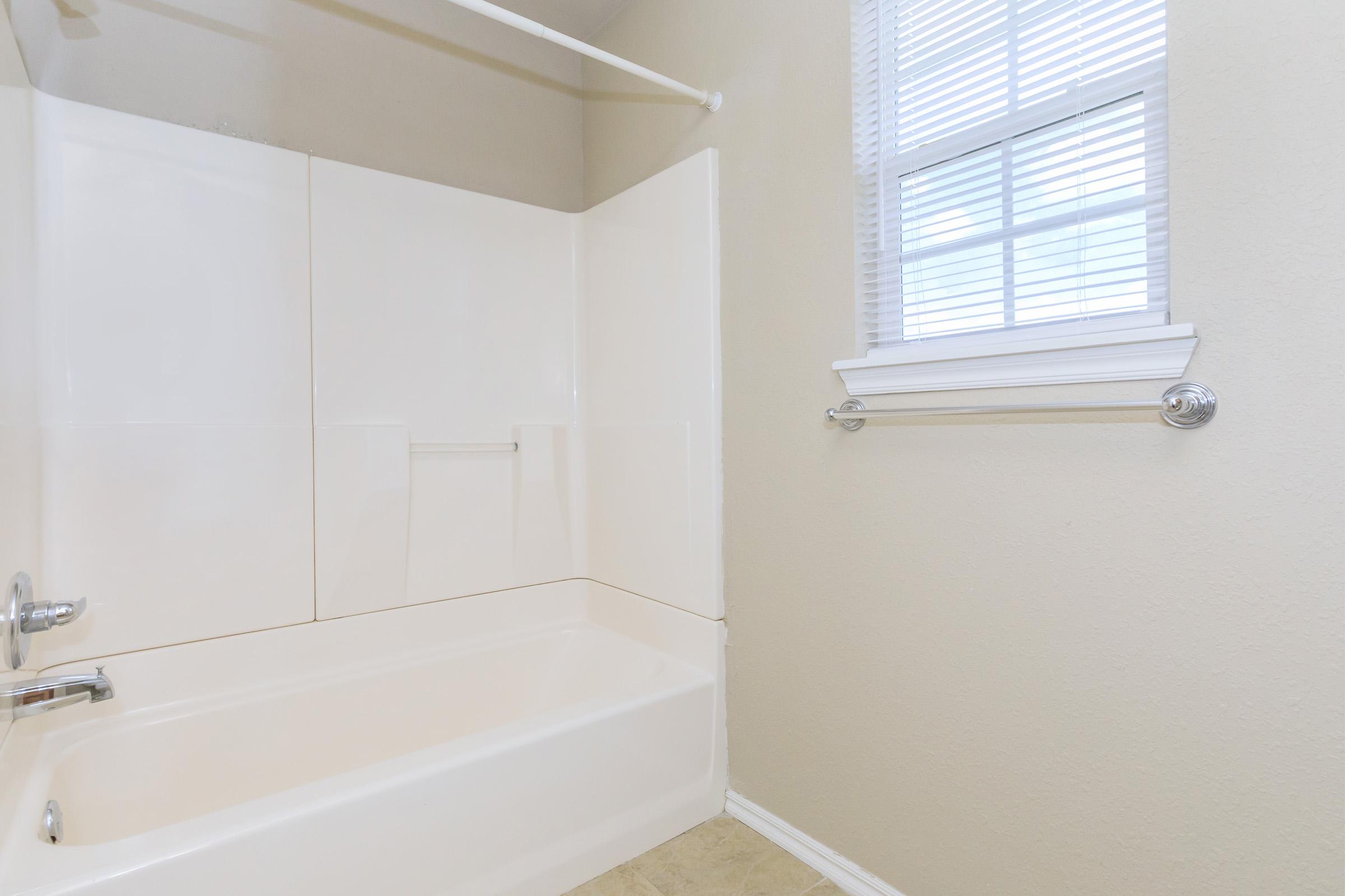 Bright and clean bathroom featuring a bathtub with a showerhead, white walls, and a window with blinds for natural light. The floor is tiled, and there's a towel rack mounted next to the window.