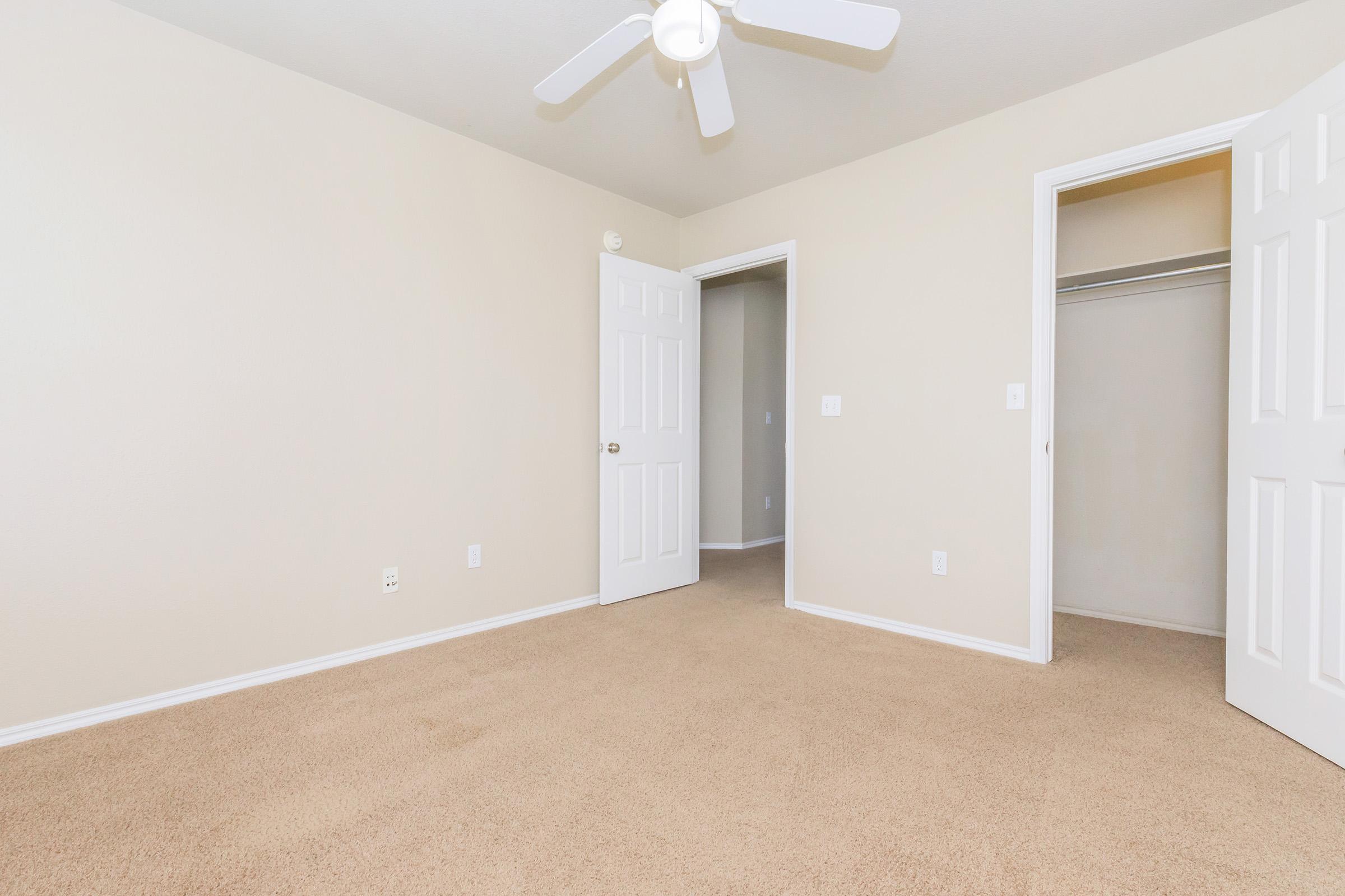 A clean, empty bedroom with light beige walls and plush beige carpeting. The room features a ceiling fan and two doors, one leading to a closet and the other to a hallway. Natural light brightens the space, creating a tranquil atmosphere.