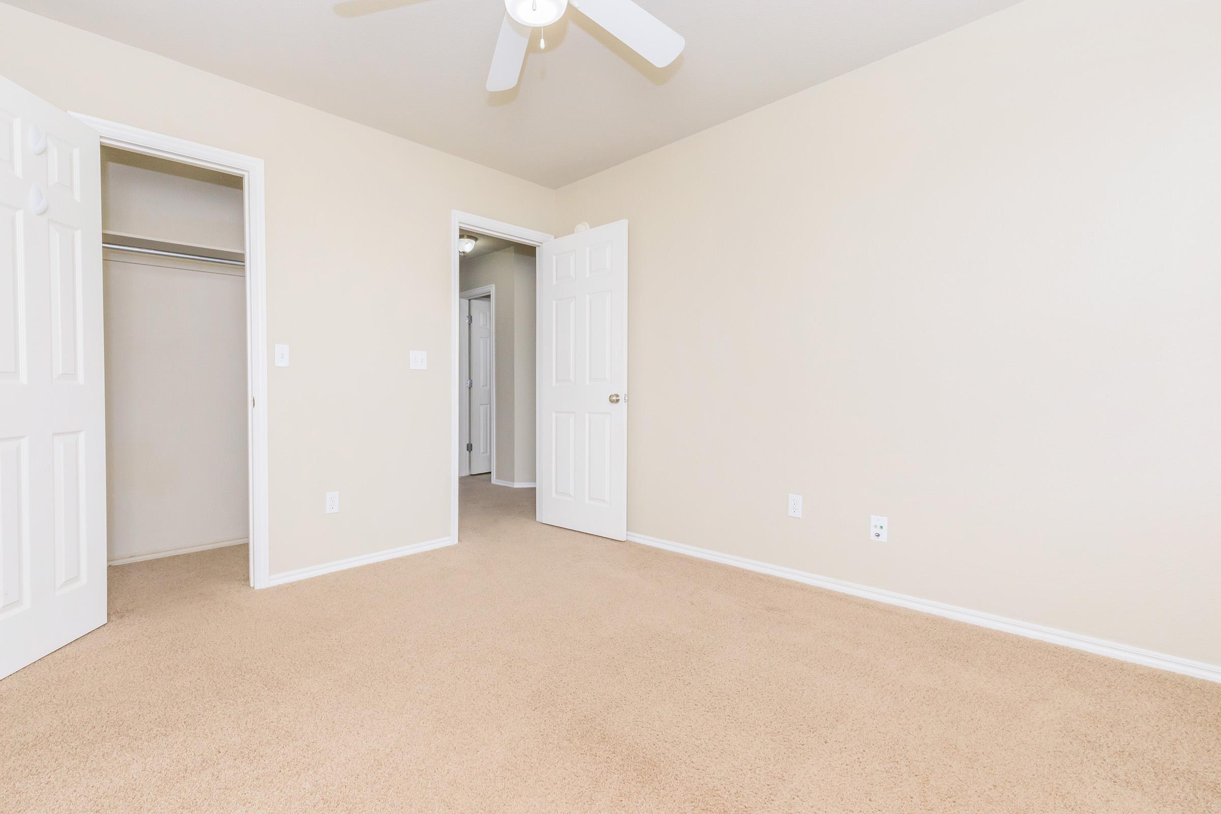 Empty room with light beige walls and carpet, featuring a ceiling fan and two open white doors leading to a closet and another space. The room is well-lit, showcasing a minimalist design.