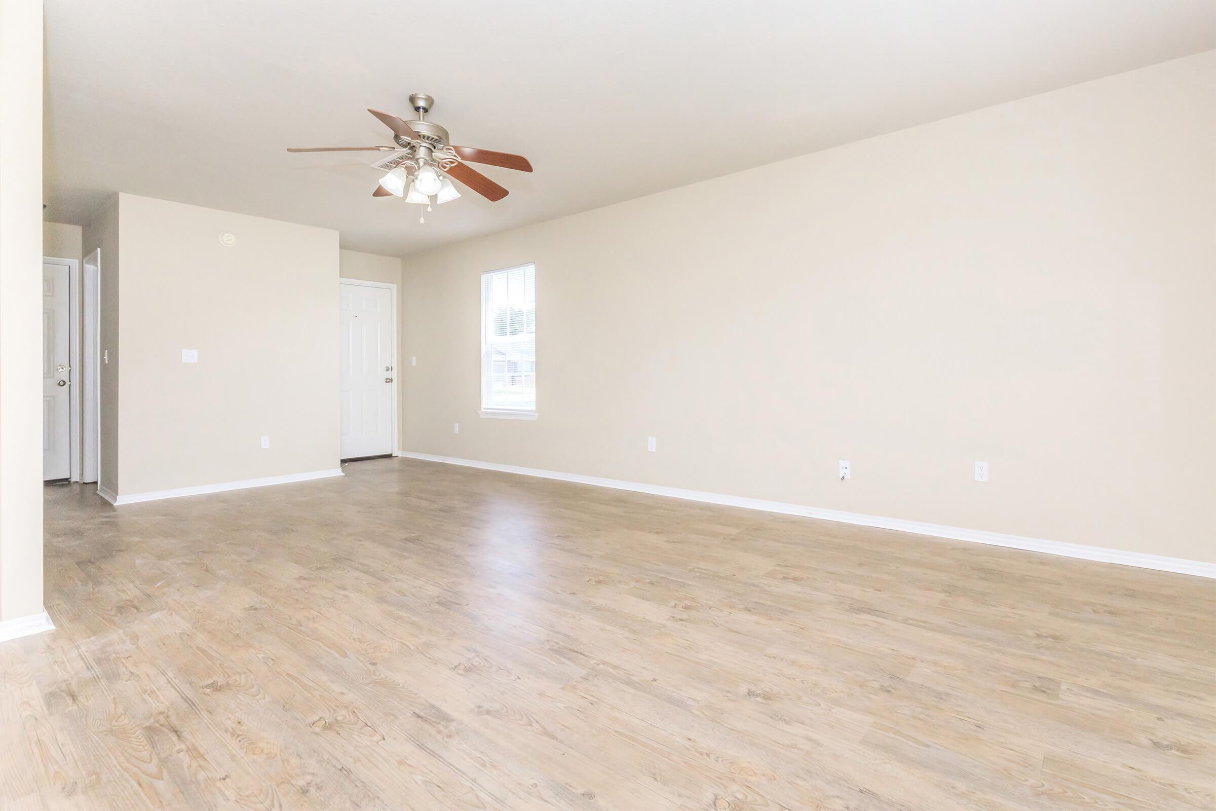 A bright, empty living room with light-colored walls and a wood-style floor. A ceiling fan with wooden blades is visible, and there's a window allowing natural light. To the left, a door leads outside, while another door is slightly open, indicating access to a hallway.