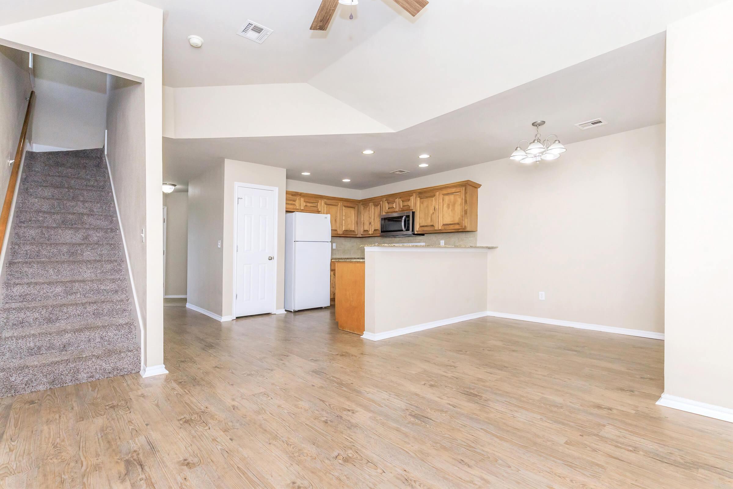 A spacious, open-concept living area with light-colored walls and wooden flooring. To the left, there is a staircase leading to another level. The kitchen features wooden cabinets, a white refrigerator, and a microwave. Natural light fills the room, creating a welcoming atmosphere.