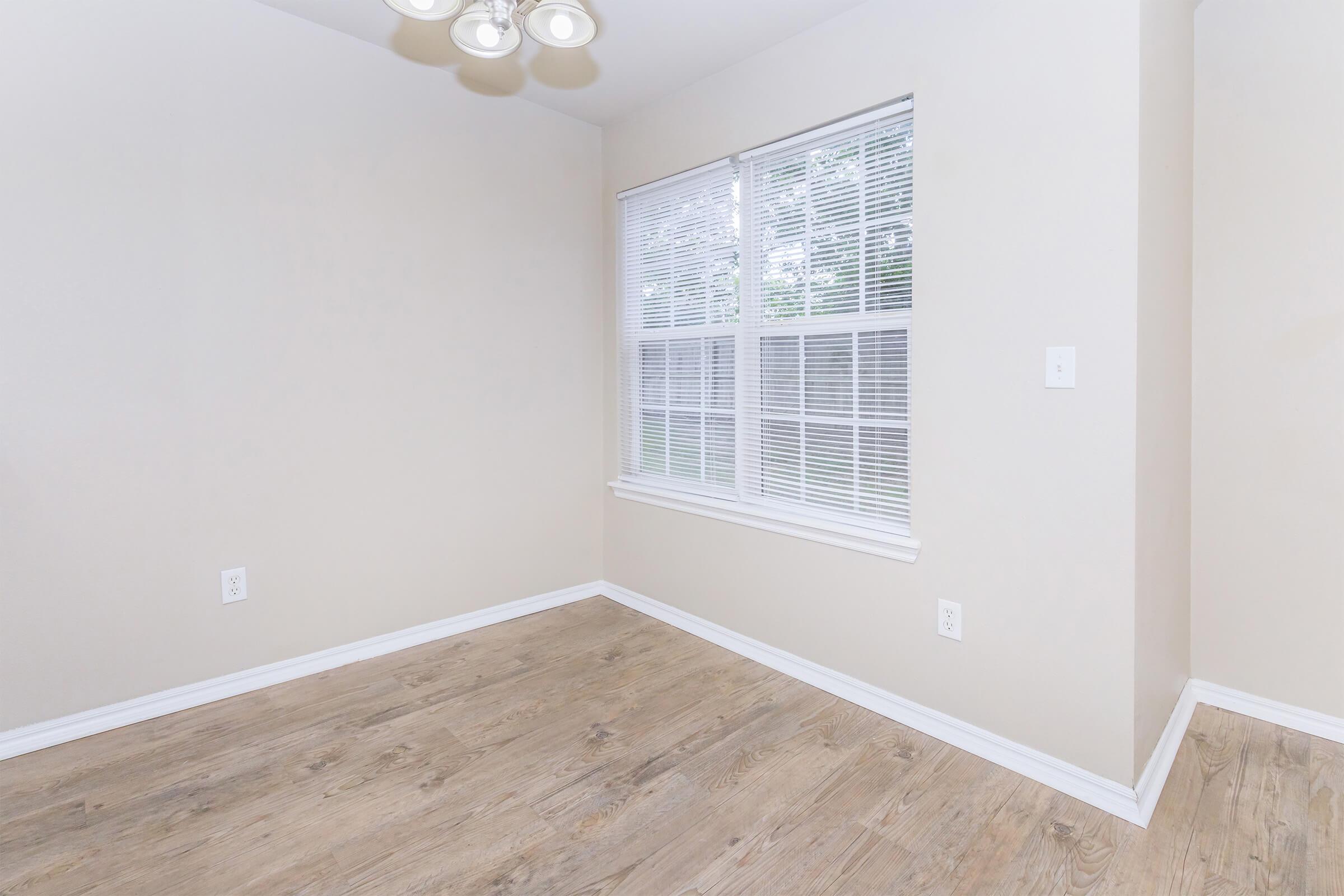 Empty room with light beige walls and hardwood flooring. A large window with white blinds lets in natural light, and a modern light fixture hangs from the ceiling. The space is devoid of furniture, highlighting its bright, airy feel.