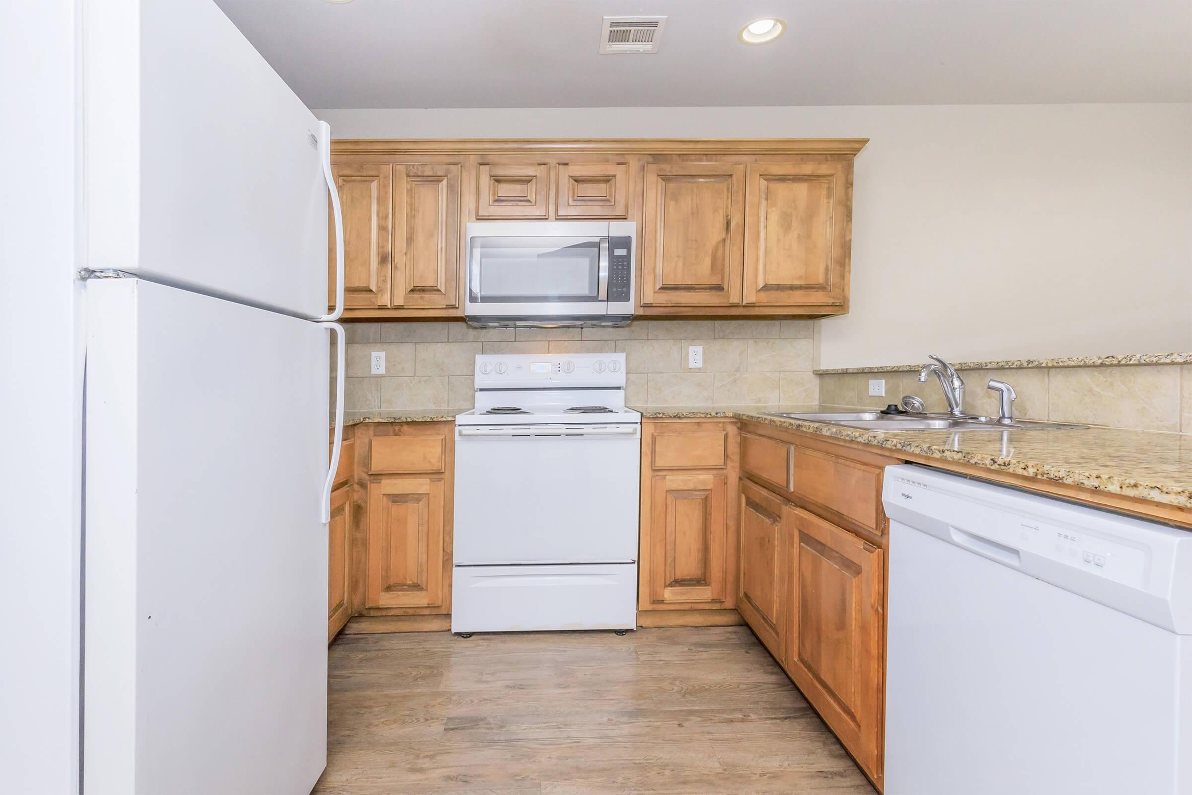 Modern kitchen featuring light wooden cabinets, a white stove, and a microwave above. A stainless steel sink is situated next to a white dishwasher and refrigerator. The countertops are made of granite, and the flooring is a light wood finish. The overall design is clean and functional.
