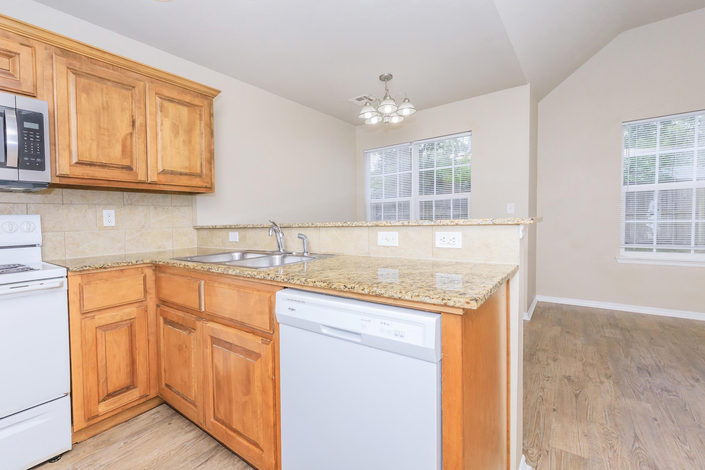 A bright kitchen featuring wooden cabinets, a granite countertop, a white dishwasher, and a gas stove. Natural light comes through two windows in the adjacent living area, showcasing a spacious layout with wooden flooring. The walls are painted in a neutral color, creating a warm and inviting atmosphere.