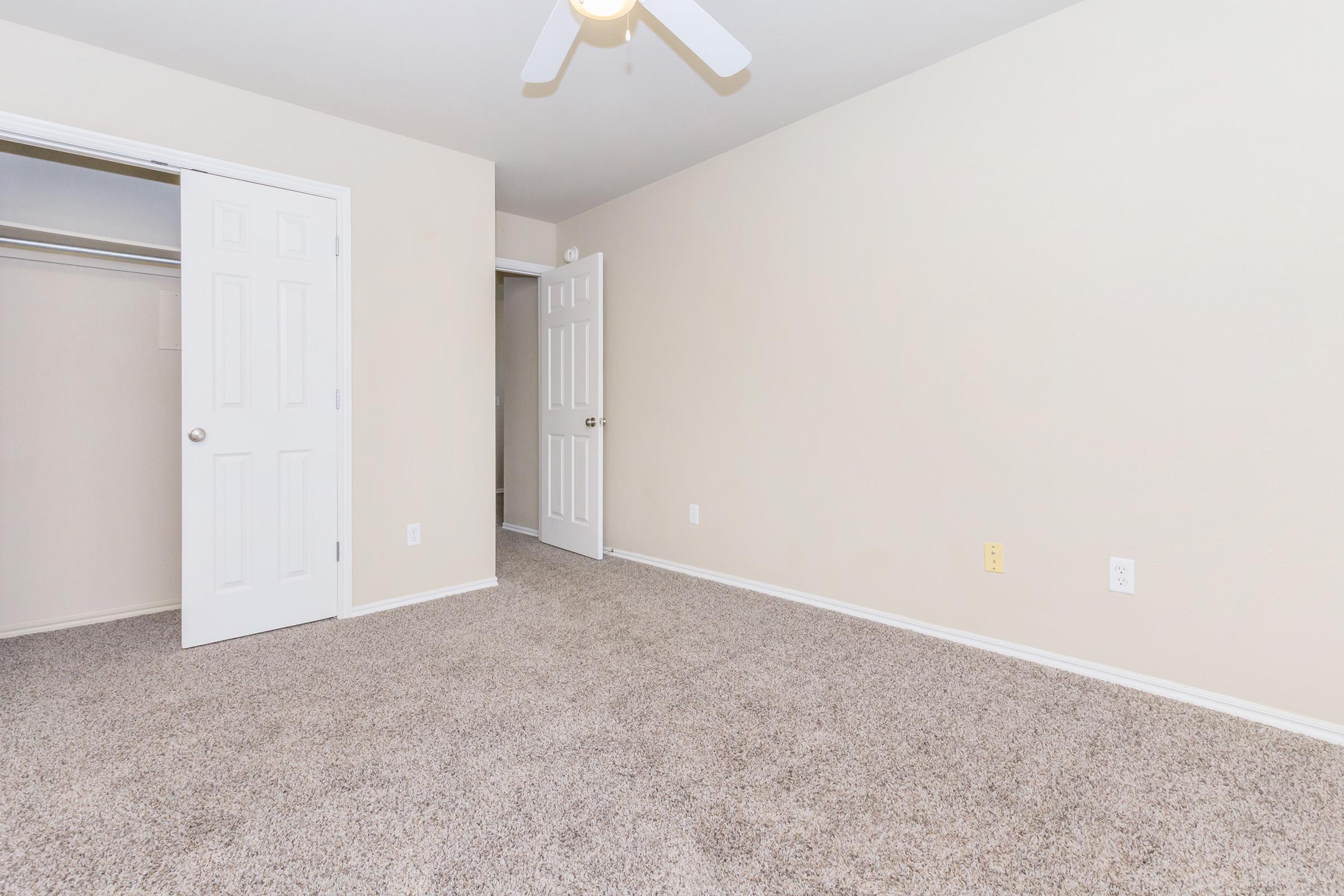 A spacious, empty room with light beige walls and plush carpet. Two closed white doors are visible: one leads to a closet, and the other opens to another room. A ceiling fan hangs from the ceiling, offering a modern touch to the minimalist design. The room is well-lit and inviting.