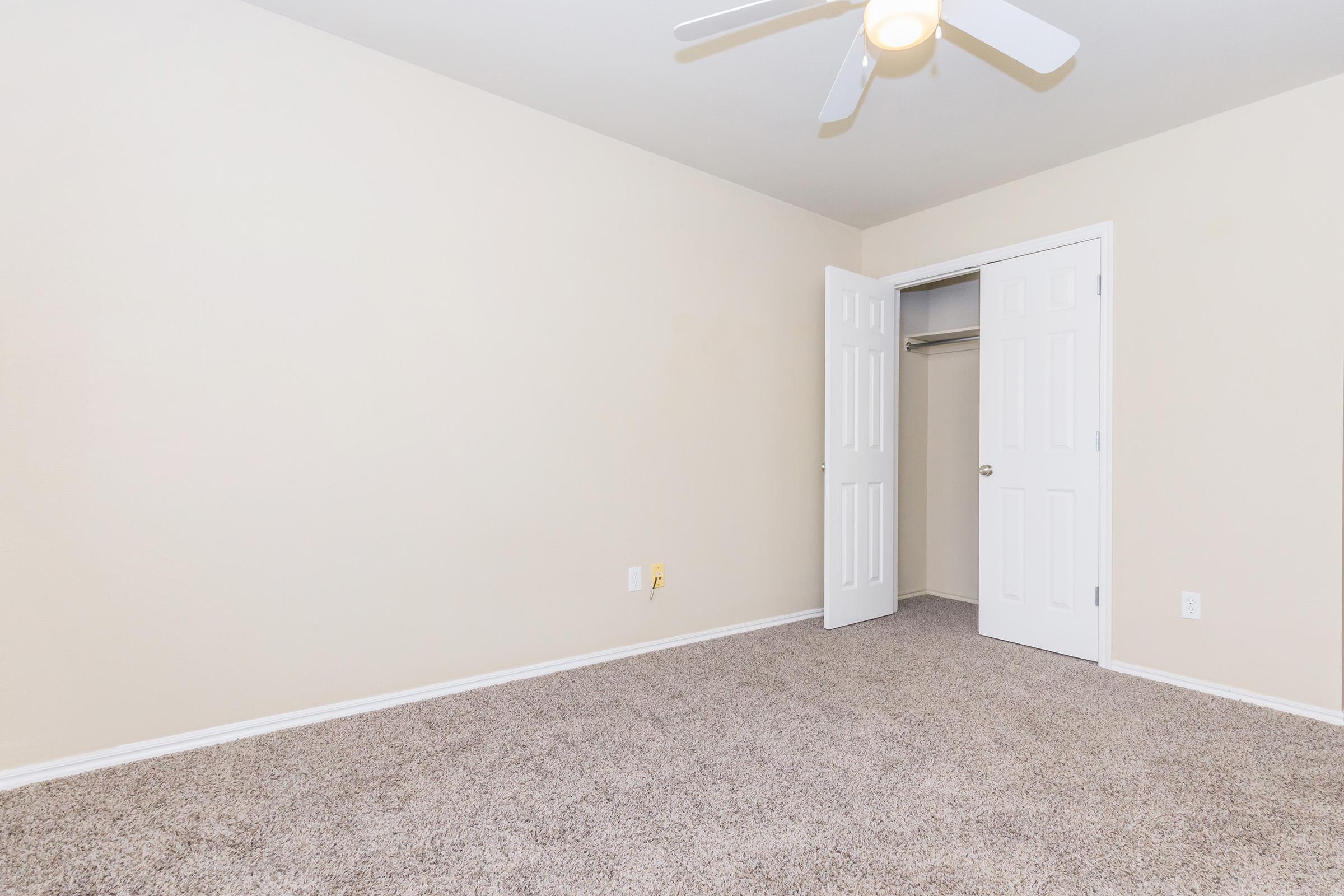 An empty room with beige walls and carpeted floor. There are two white doors, one of which leads to a closet. A ceiling fan is visible, and the room is well-lit with natural light. The space appears clean and ready for furniture.