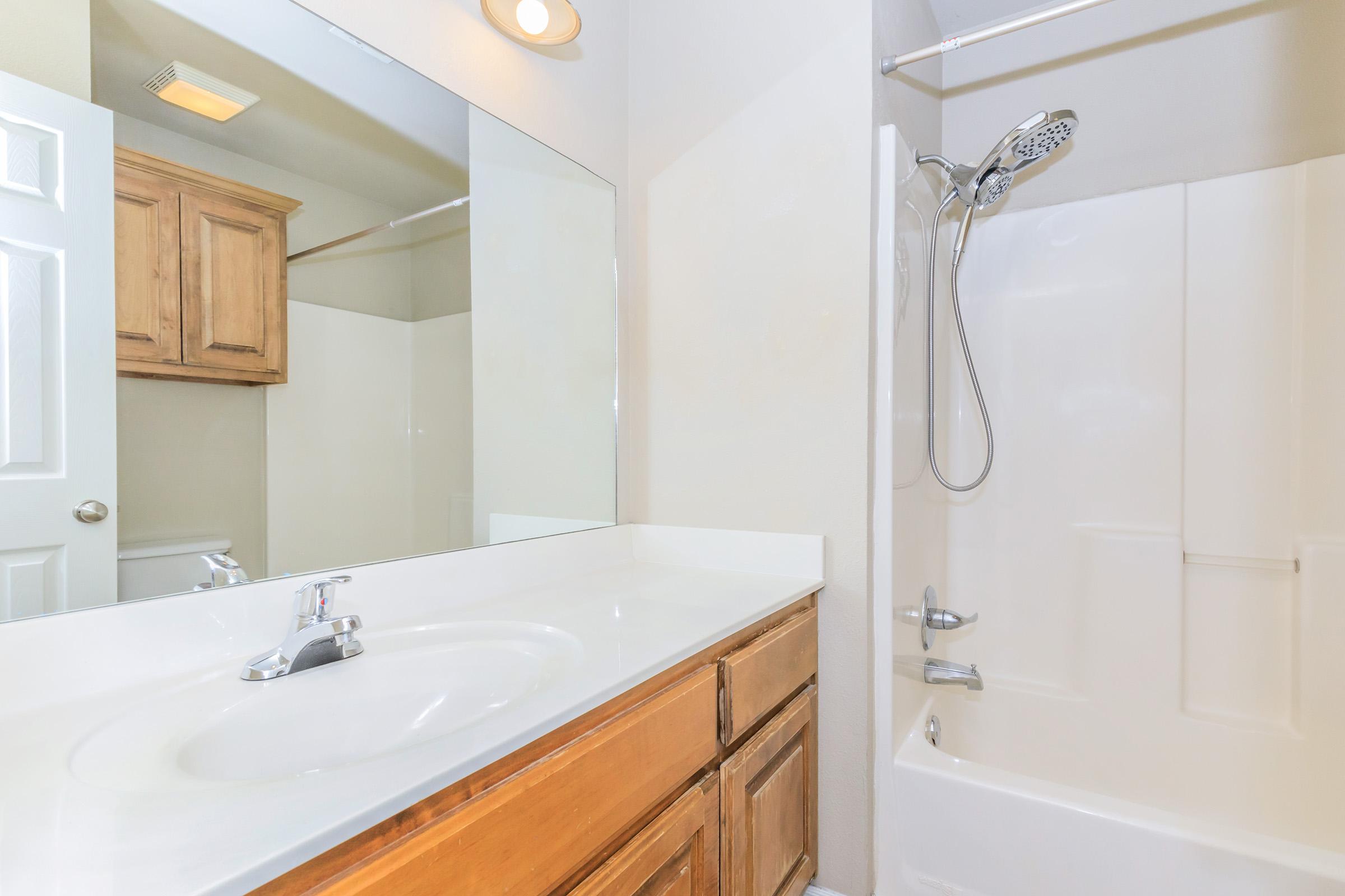 A clean bathroom featuring a white sink and countertop, wooden cabinets, and a shower with a sliding glass door. A large mirror reflects the space, and the walls are painted in a light color, giving a bright and spacious feel.