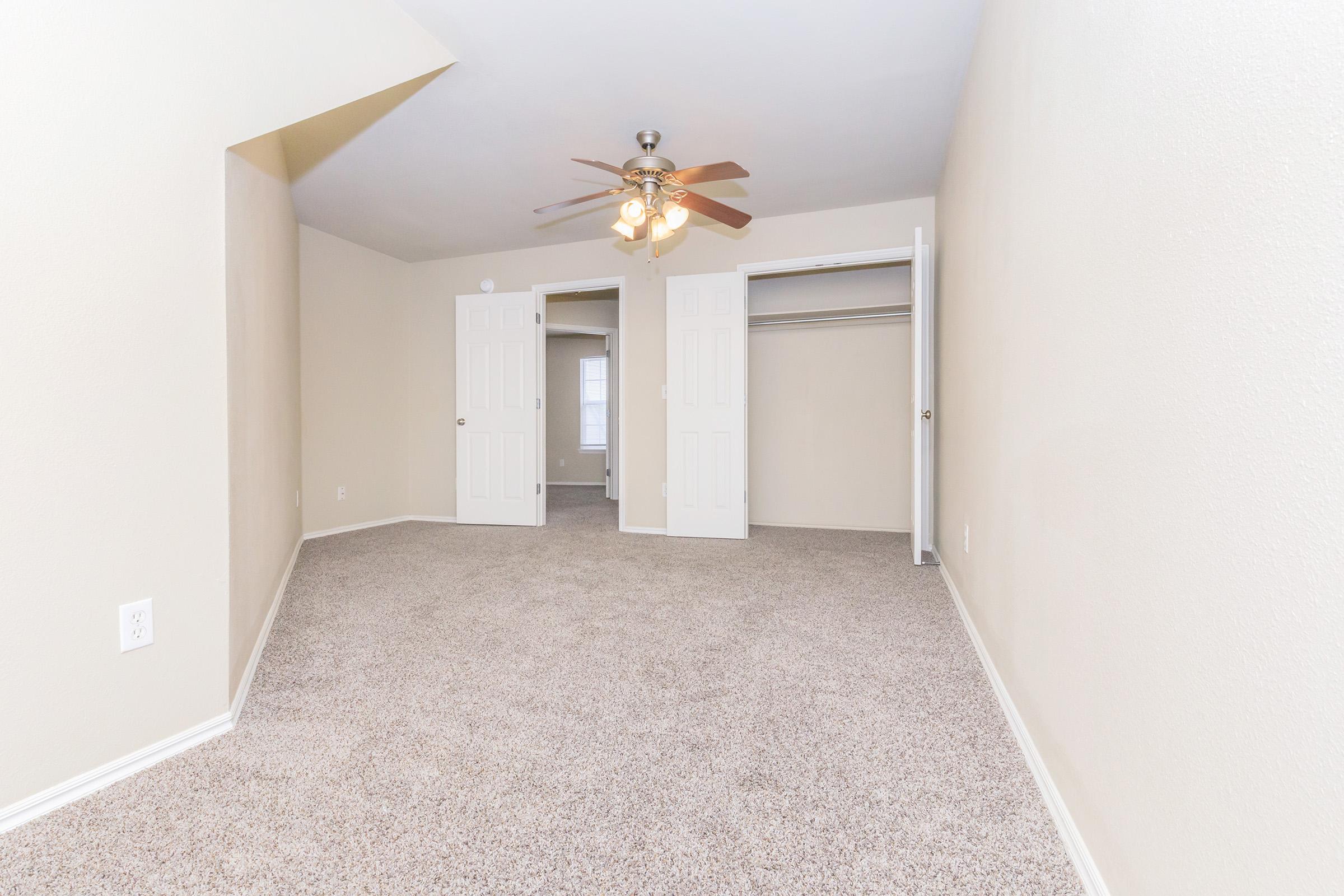 A spacious room with beige walls and light carpet, featuring a ceiling fan with three bulbs. There are two white closet doors on the left and another door leading to a small hallway in the background. Natural light enters the room, creating a bright and inviting atmosphere.