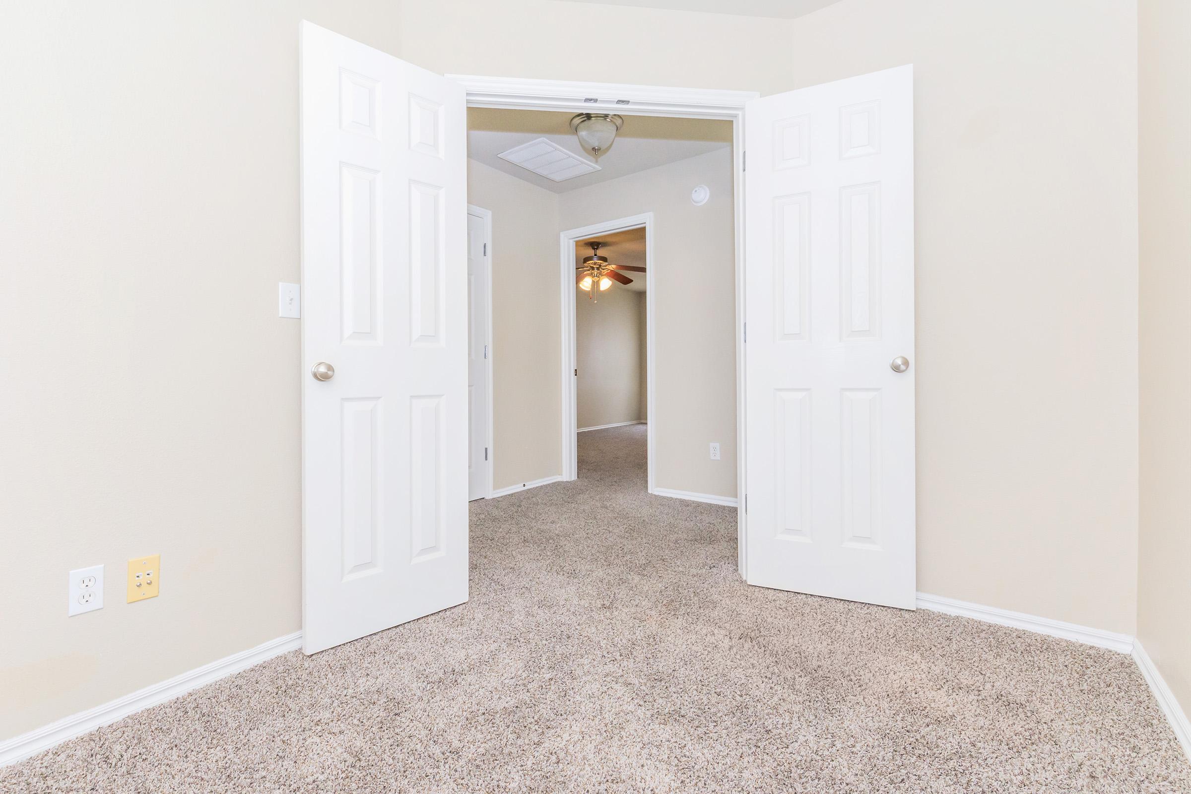 A spacious, neutral-colored room seen through open double doors. The room features beige walls and carpet, with a ceiling fan visible in the background. The doorway leads to another area, creating an inviting and open atmosphere. Bright lighting enhances the overall feel of the space.
