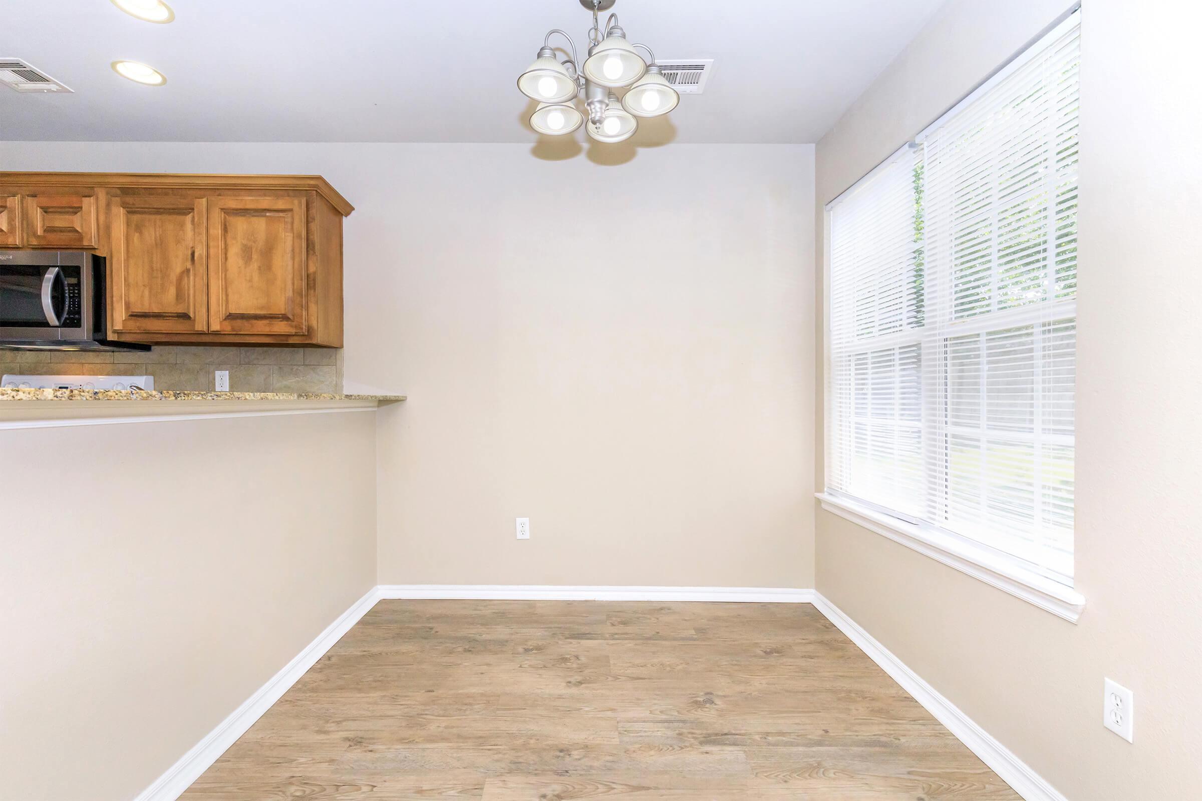 An empty room featuring light-colored walls, a wood-like floor, and a kitchen area with wooden cabinets. A modern chandelier hangs from the ceiling, and there's a large window with white blinds allowing natural light to enter. The space is bright and uncluttered, ideal for various uses.