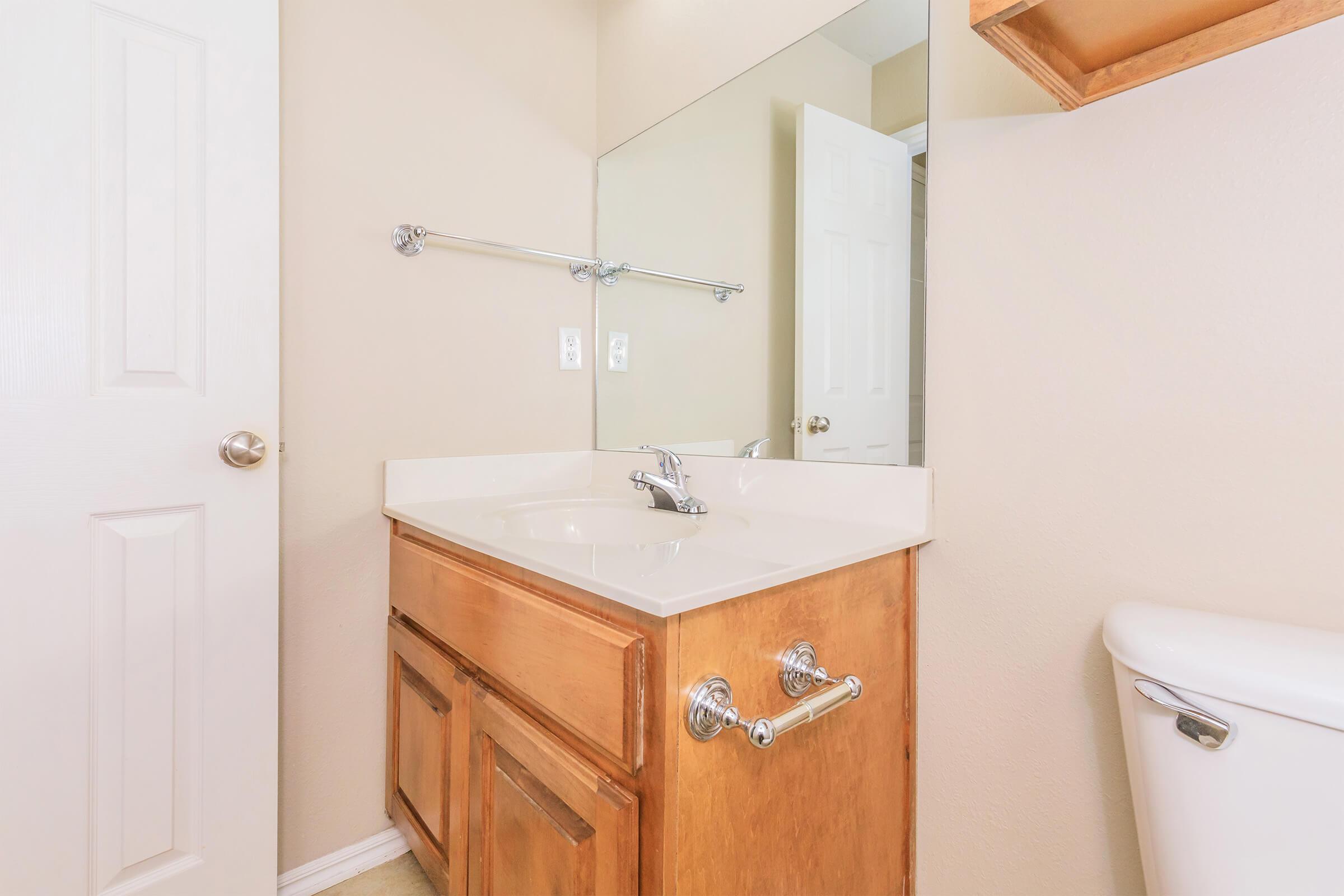 A bathroom featuring a wooden vanity with a sink and faucet, a large mirror above the sink, a toilet to the right, and a door on the left side. The walls are painted in a neutral color, and the floor is light-colored. The space appears clean and well-lit.