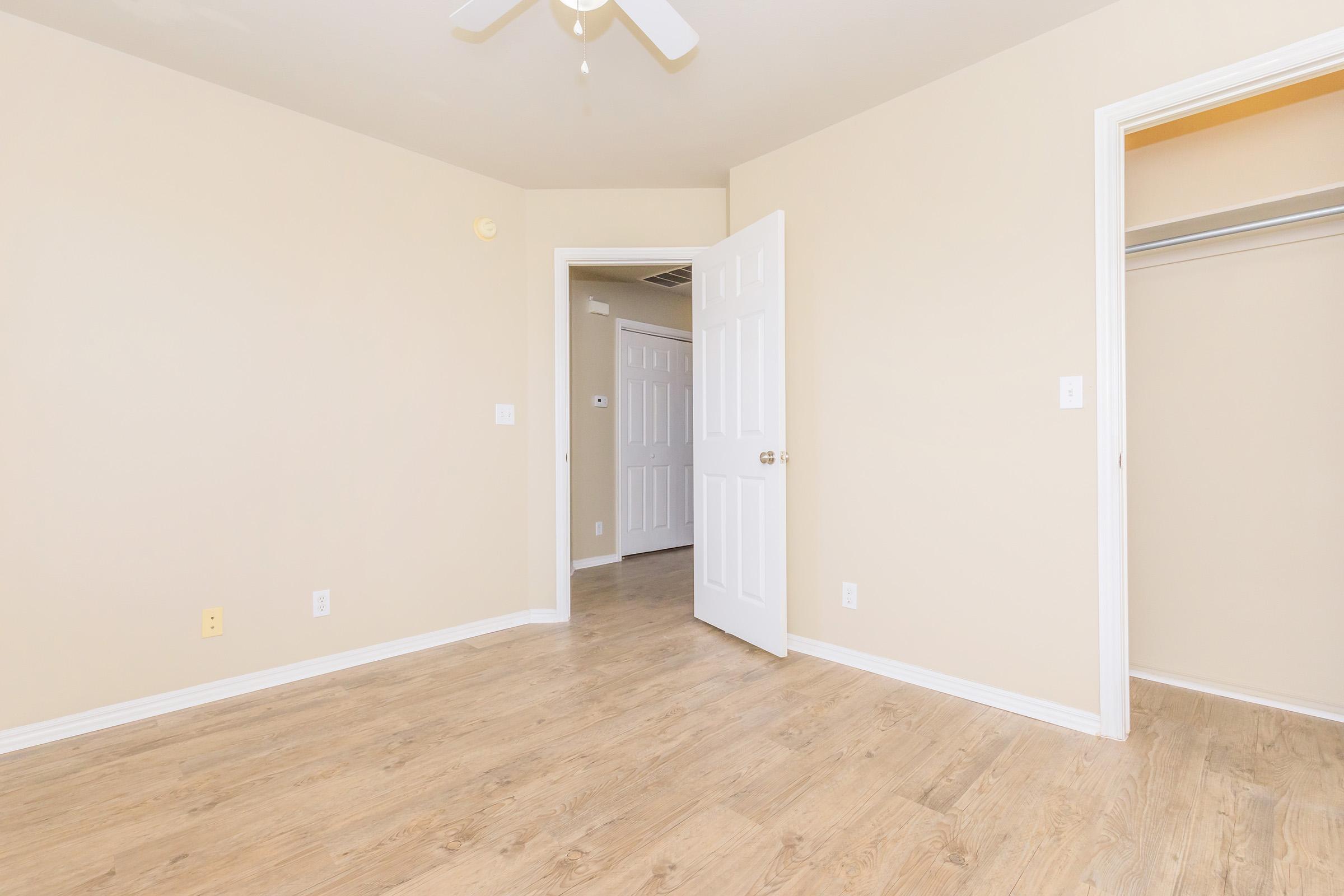 A well-lit, empty room with beige walls and wood-style flooring. There are two doors: one leading to a closet on the right and another to an adjacent room. A ceiling fan is visible, and the overall space appears inviting and clean, suitable for various uses.