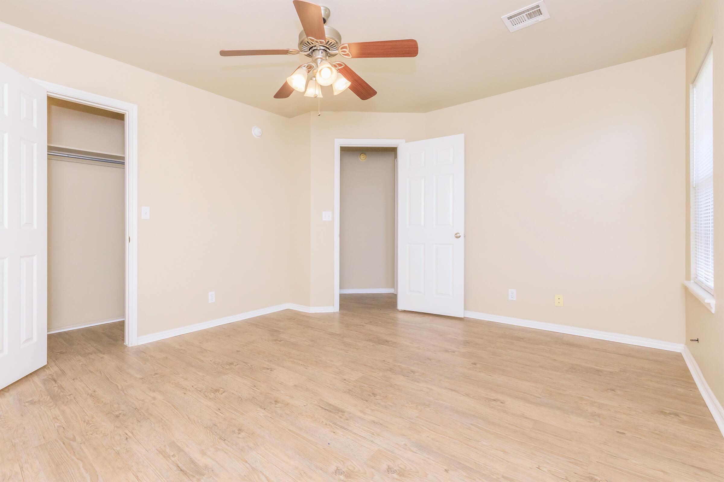 A spacious, empty room with light beige walls and wooden flooring. There is a ceiling fan with lights, two doors leading to closets, and a window allowing natural light. The room's neutral decor suggests it is ready for furnishing or decorating.