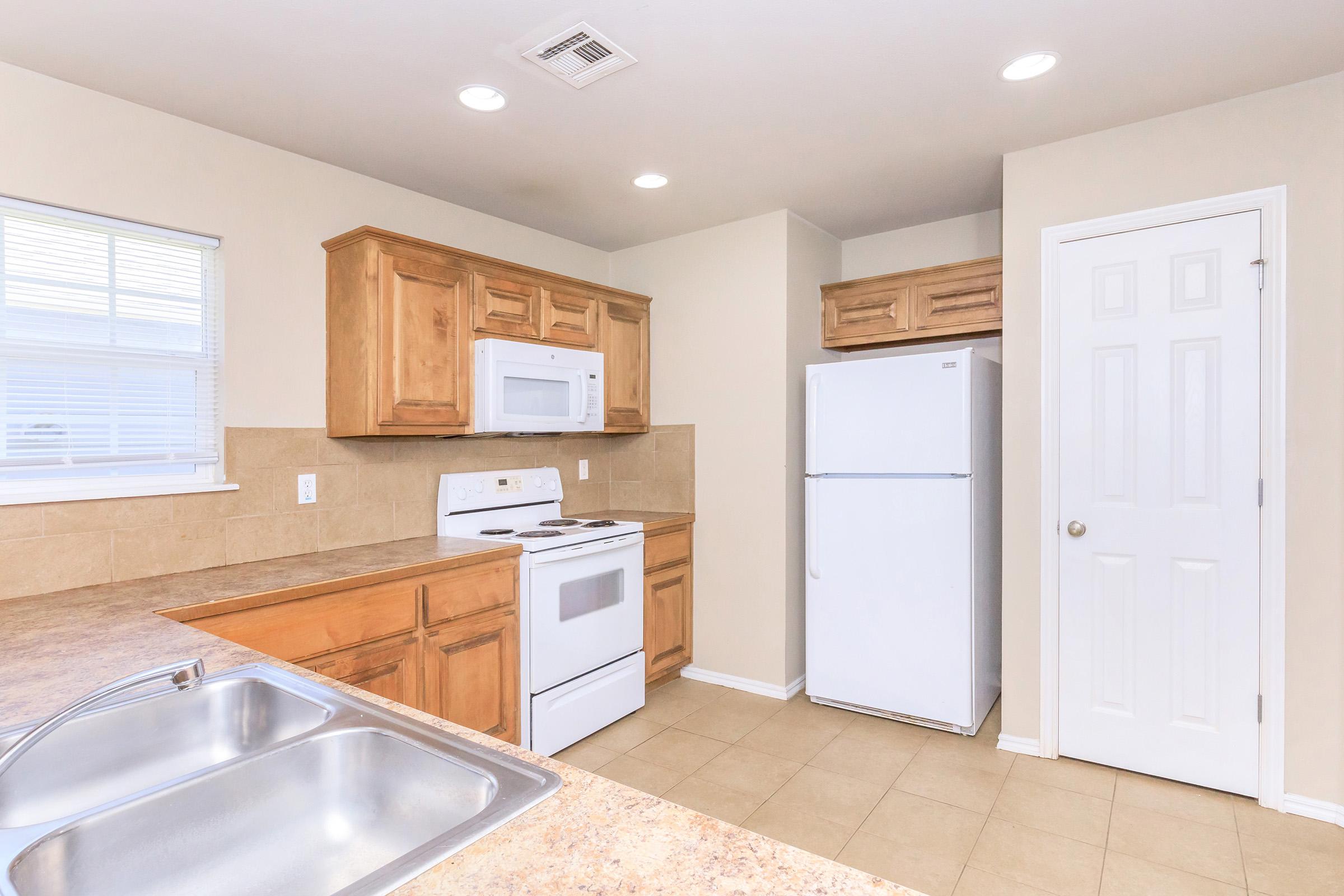A clean and modern kitchen featuring light wood cabinetry, a white refrigerator, an electric stove, and a microwave. The countertops are beige with a sink in the foreground. Natural light comes in through a window, and the walls are painted a soft neutral color.