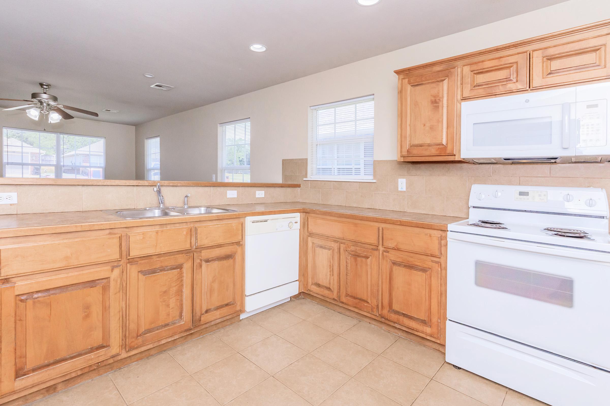 Modern kitchen featuring light wood cabinetry, a double sink, a dishwasher, and a white stove. The countertops are tiled, and there are large windows allowing natural light. The flooring is also tiled, contributing to a clean and spacious appearance.