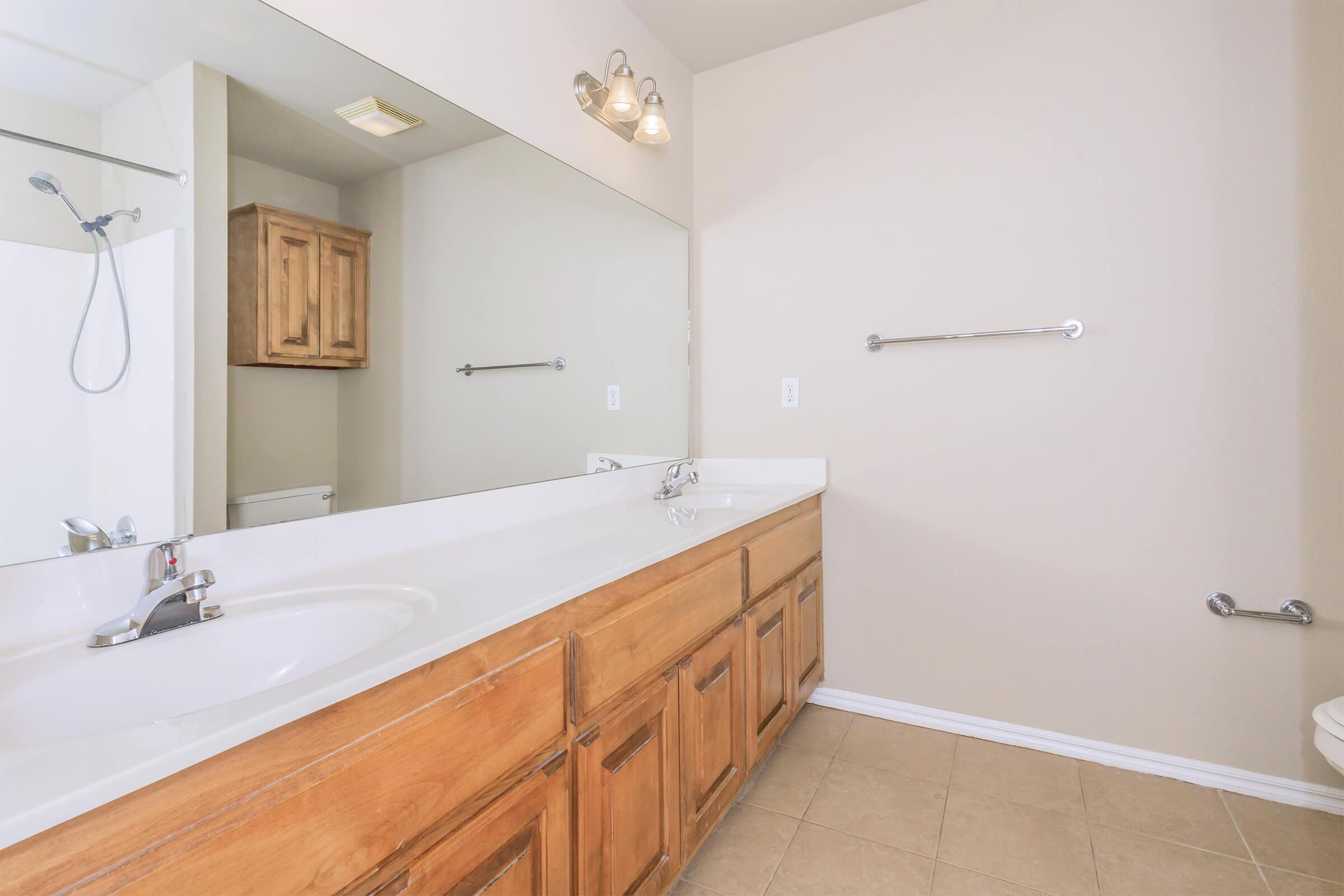 A spacious bathroom featuring a large mirror above a double sink with wooden cabinetry. The light-colored walls are complemented by bright lighting fixtures. A shower with a glass door is visible in the corner, along with a toilet. The floor is tiled, adding a clean and modern look to the space.