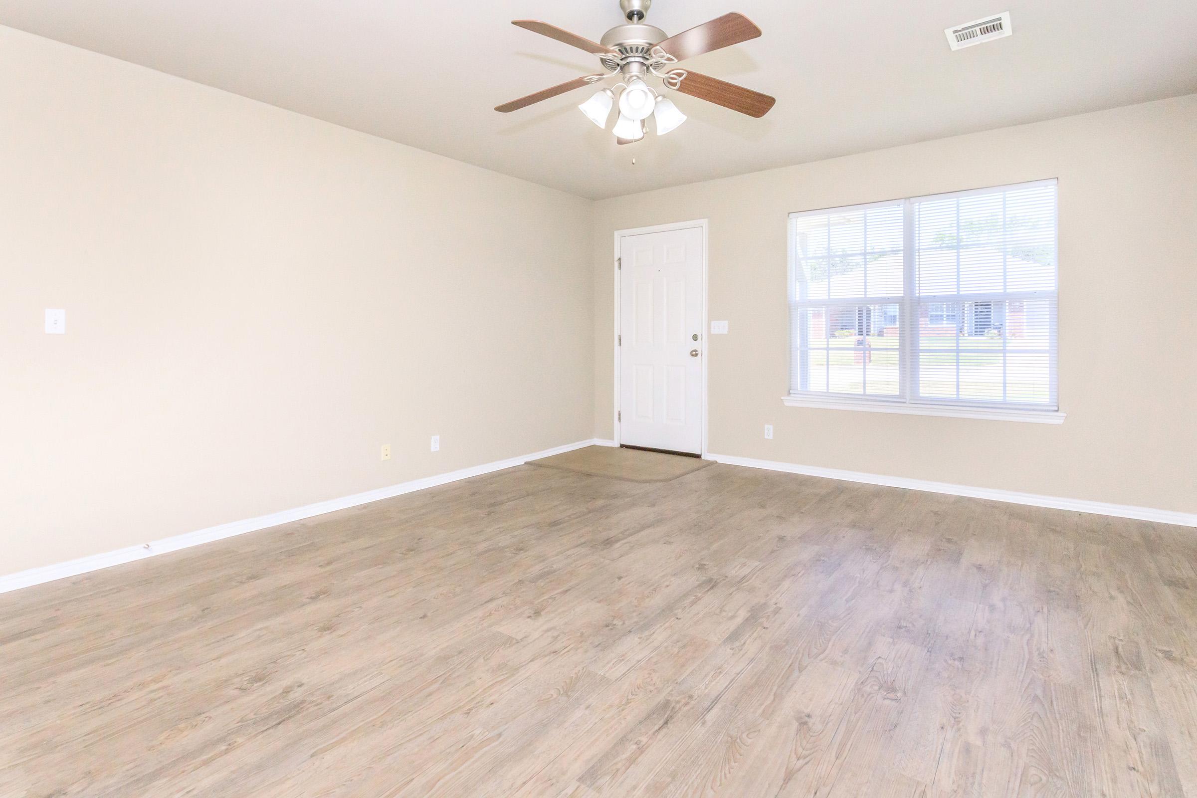An empty living room featuring light-colored walls, a light wood floor, and a ceiling fan with wooden blades. The room has a front door on one side and a large window with vertical blinds allowing natural light to enter.