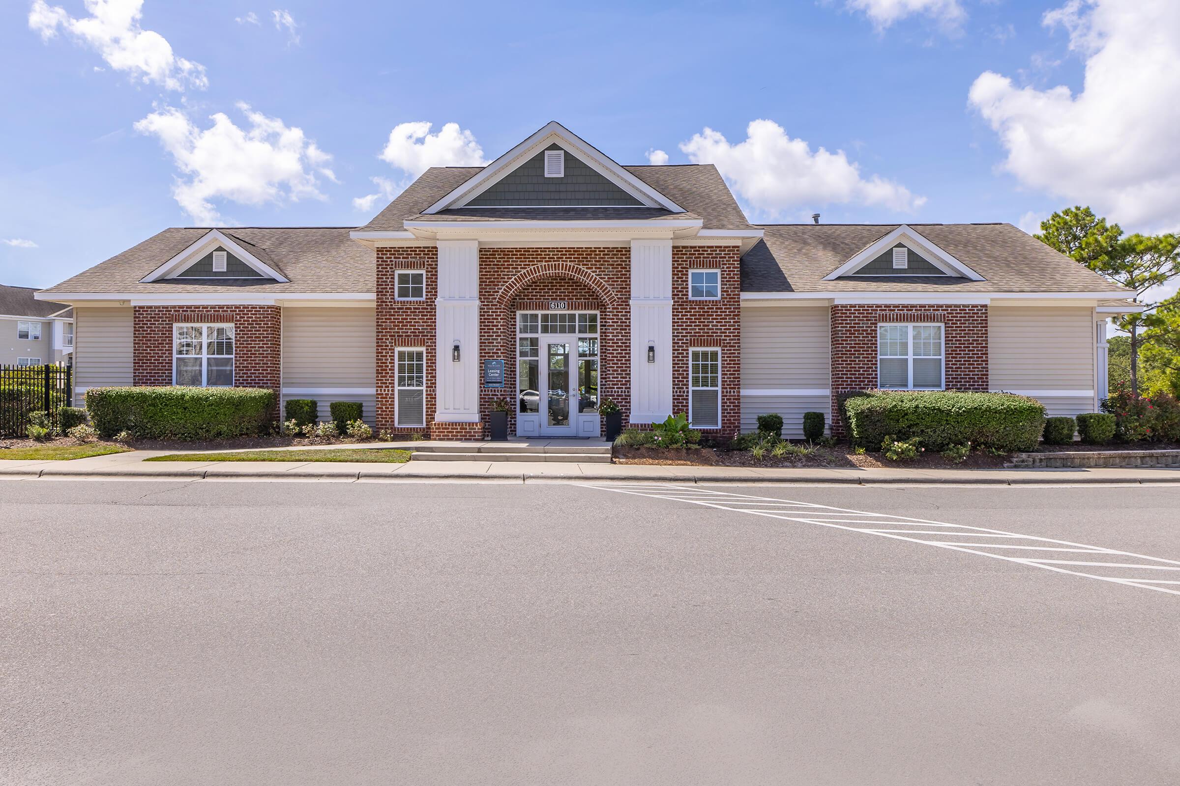 A one-story brick building with a white and gray facade, featuring a central entrance with large windows and an arched doorway. The surrounding area is landscaped with green bushes and trees, under a clear blue sky with scattered clouds.