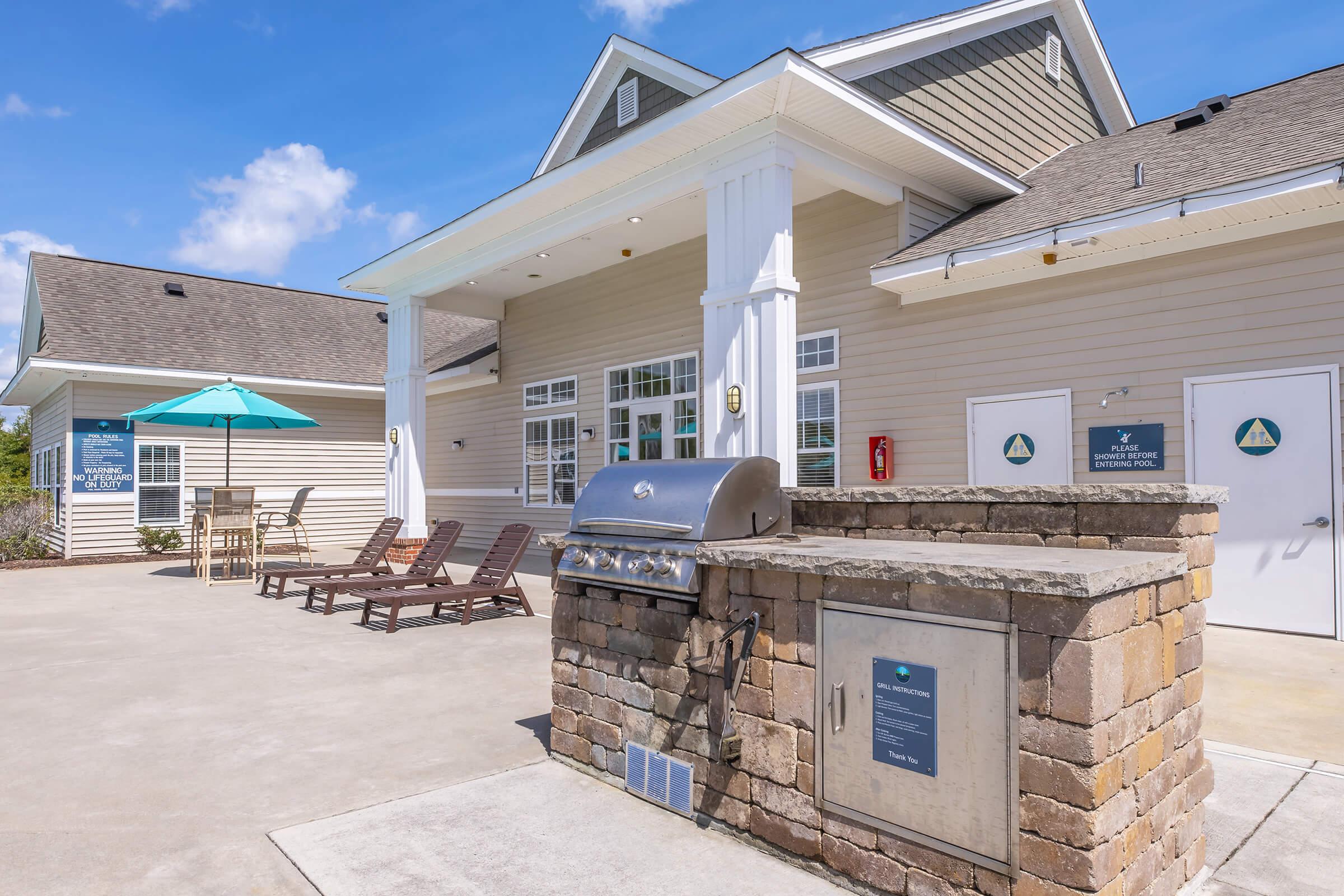 Modern outdoor area featuring a grill station with a stone enclosure, surrounded by lounge chairs and an umbrella table. In the background, a welcoming building with large windows and a porch is visible under a clear blue sky. The scene suggests a comfortable and inviting space for relaxation and social gatherings.