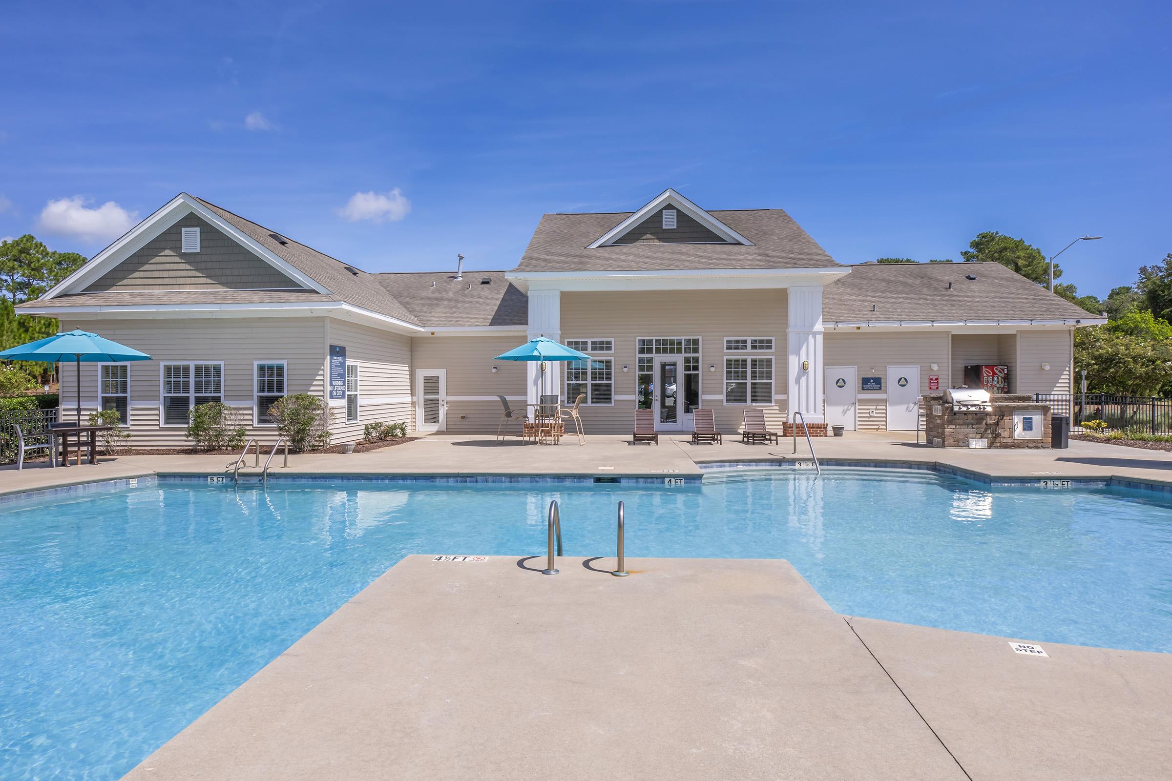 A well-maintained swimming pool with clear blue water, surrounded by lounge chairs and umbrellas. In the background, there's a stylish clubhouse with large windows and a welcoming entrance, set against a bright blue sky with a few clouds. The area is landscaped with greenery, creating a relaxing atmosphere.