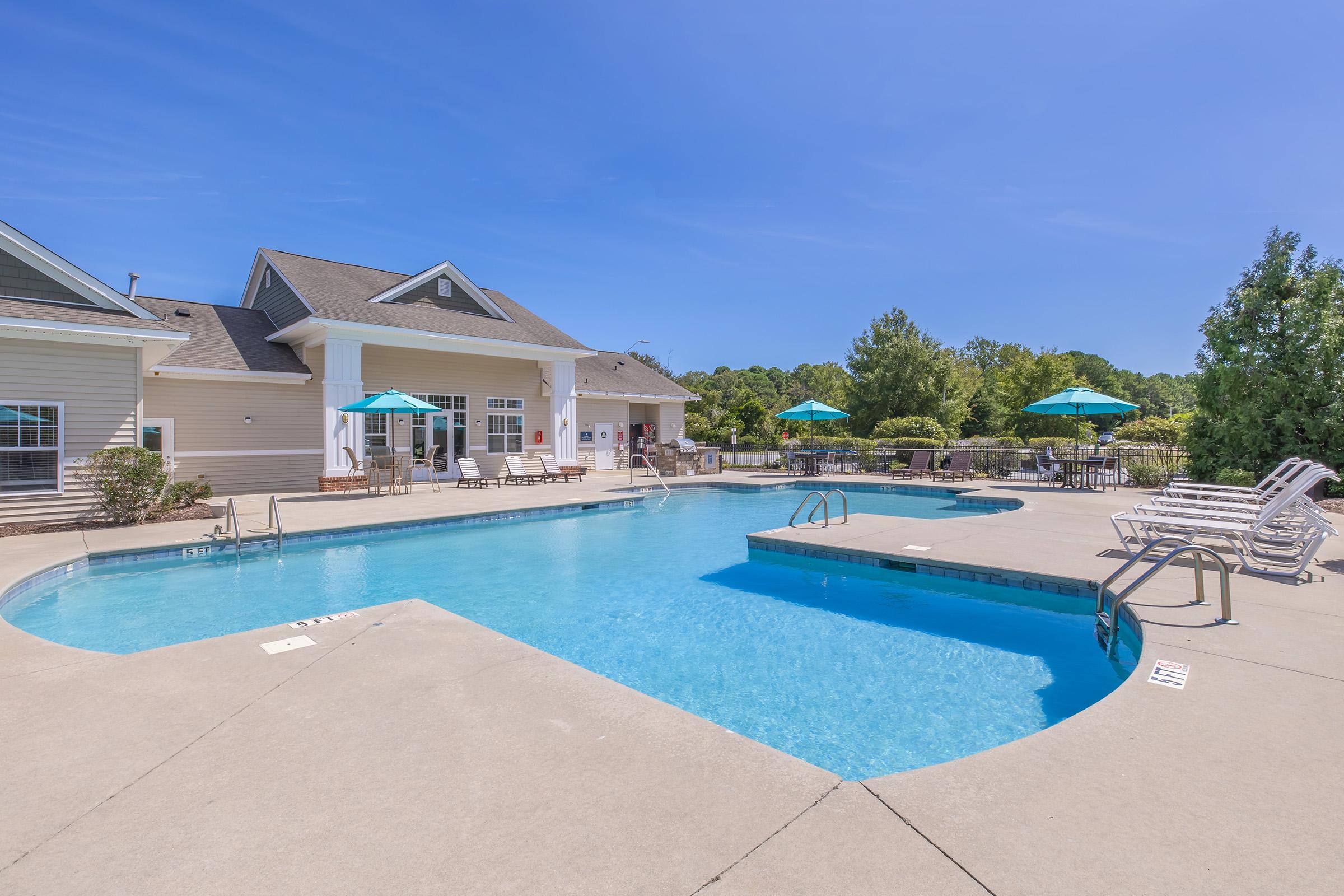 A crystal clear swimming pool with a shallow end and steps leading in. Surrounding the pool are lounge chairs and green umbrellas. In the background, there is a light-colored building and trees, all under a clear blue sky. The scene conveys a relaxing outdoor recreational area.