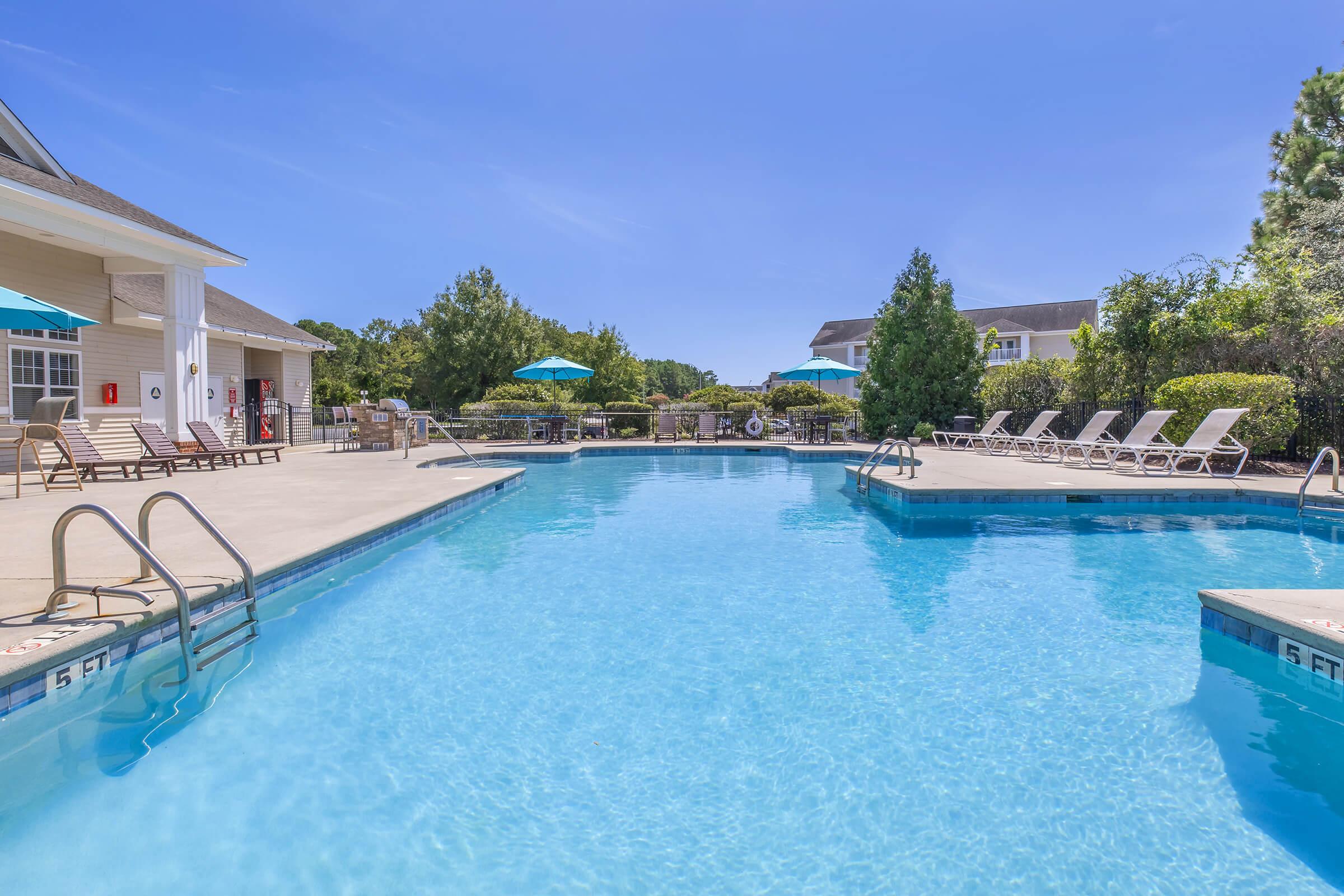 A clear blue swimming pool surrounded by lounge chairs and green trees. Umbrellas provide shade near the pool edge. The area is sunny with a bright blue sky, creating a relaxing atmosphere for leisure and recreation.