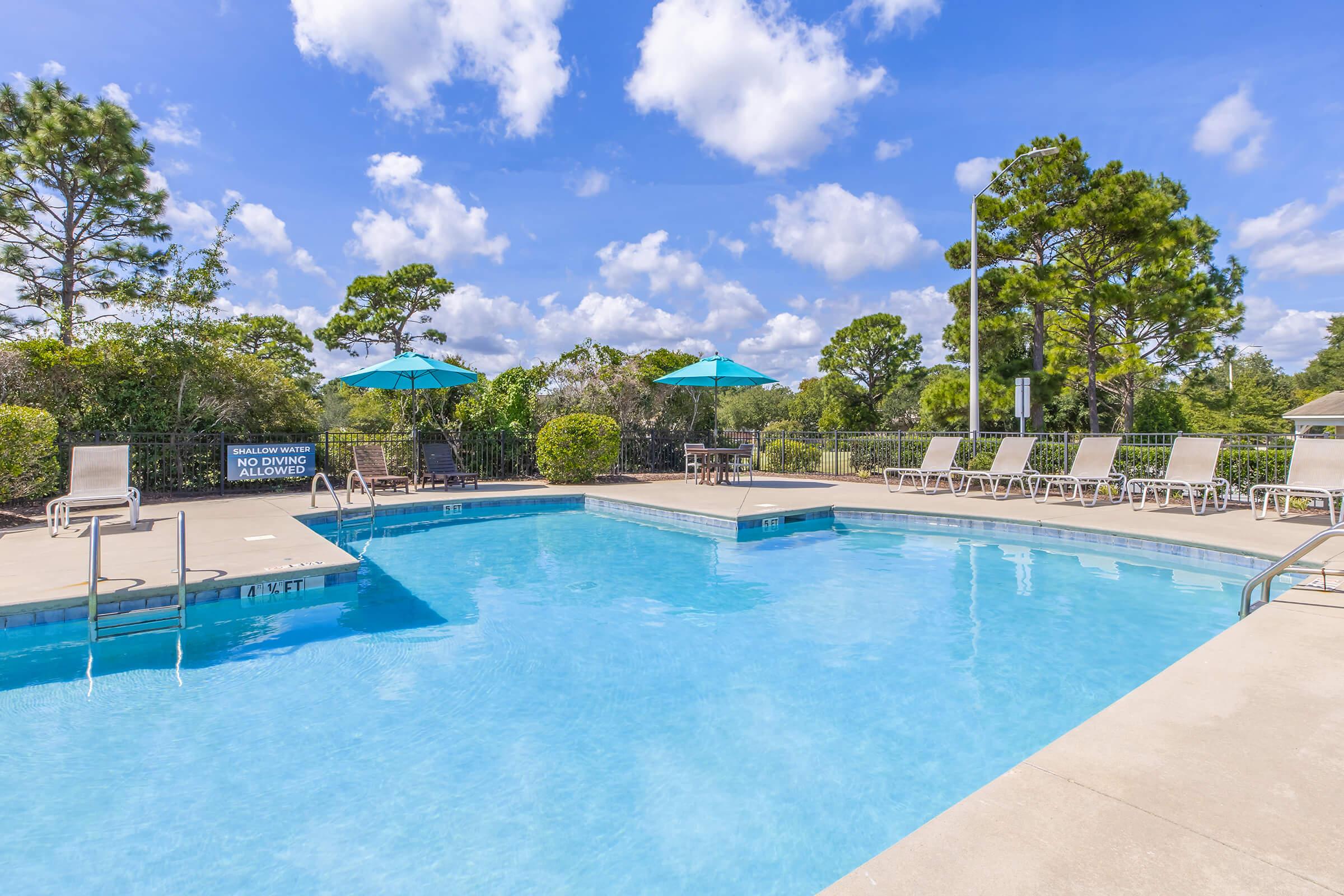 A clear blue swimming pool surrounded by lounge chairs. Umbrellas provide shade on the pool deck. Lush green trees and shrubs create a serene atmosphere, with a bright blue sky and fluffy clouds overhead.