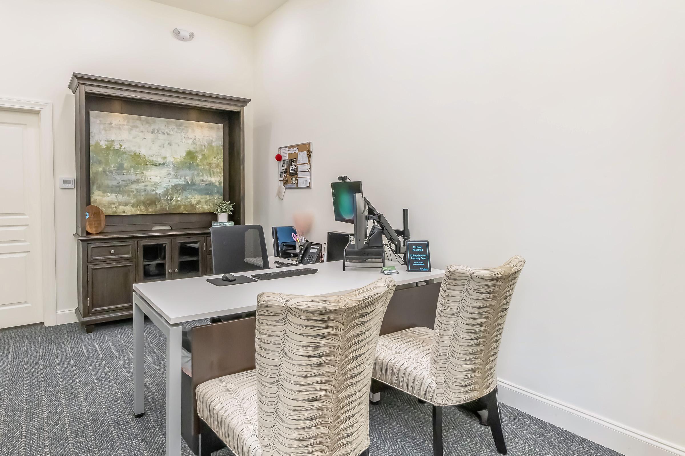 A modern office space featuring a sleek desk and two upholstered chairs. A dark wooden cabinet is against the wall, adorned with a painting and decorative items. A bulletin board and computer monitor are positioned on the desk, and the room has neutral-colored walls and a textured carpet.