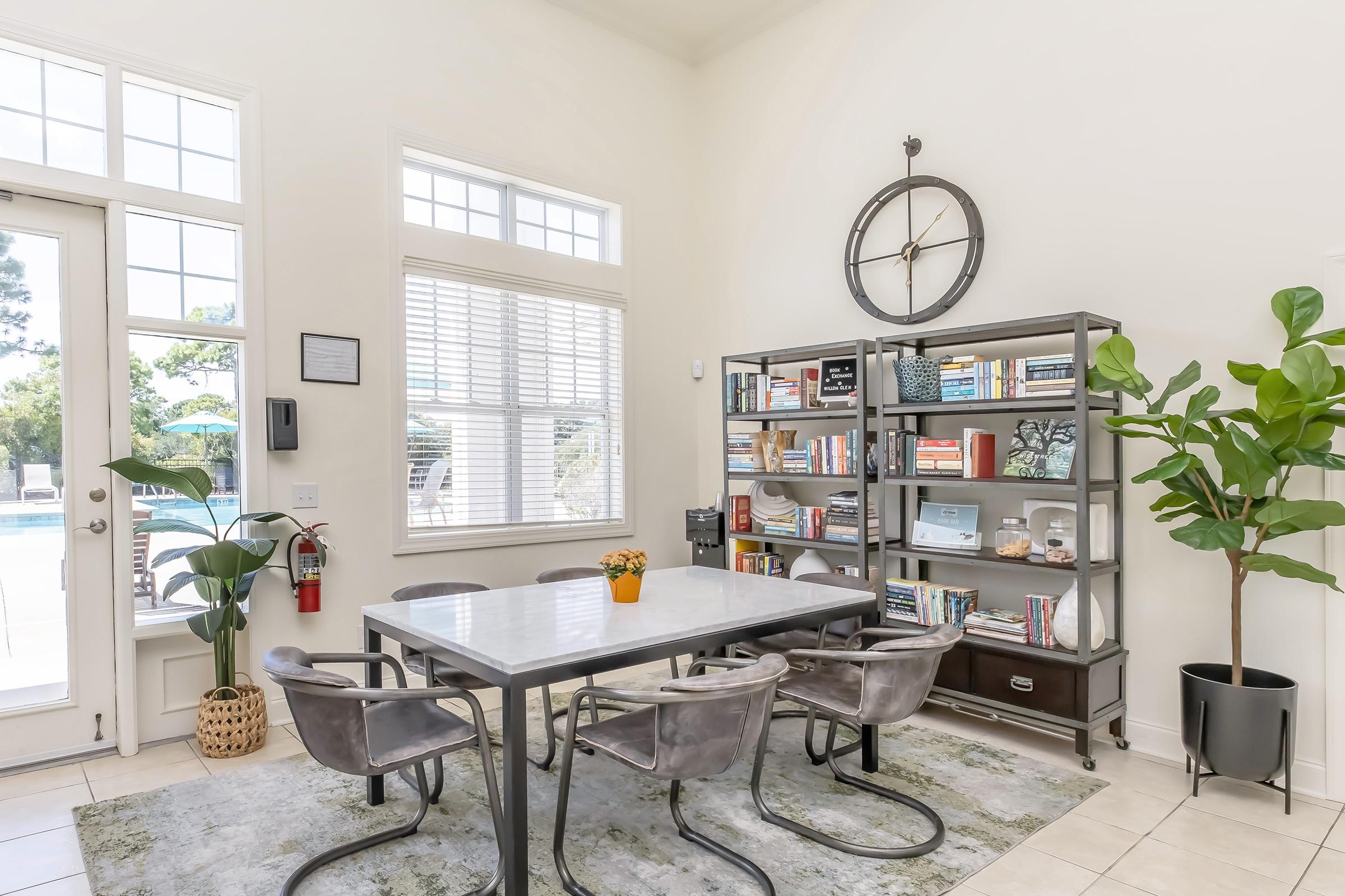A bright, modern dining area with a large table surrounded by four chairs. Against the wall, there’s a stylish bookshelf filled with books and decorative items. A potted plant adds a touch of nature, and large windows let in natural light, revealing a view of an outdoor pool. The decor is minimalistic and inviting.